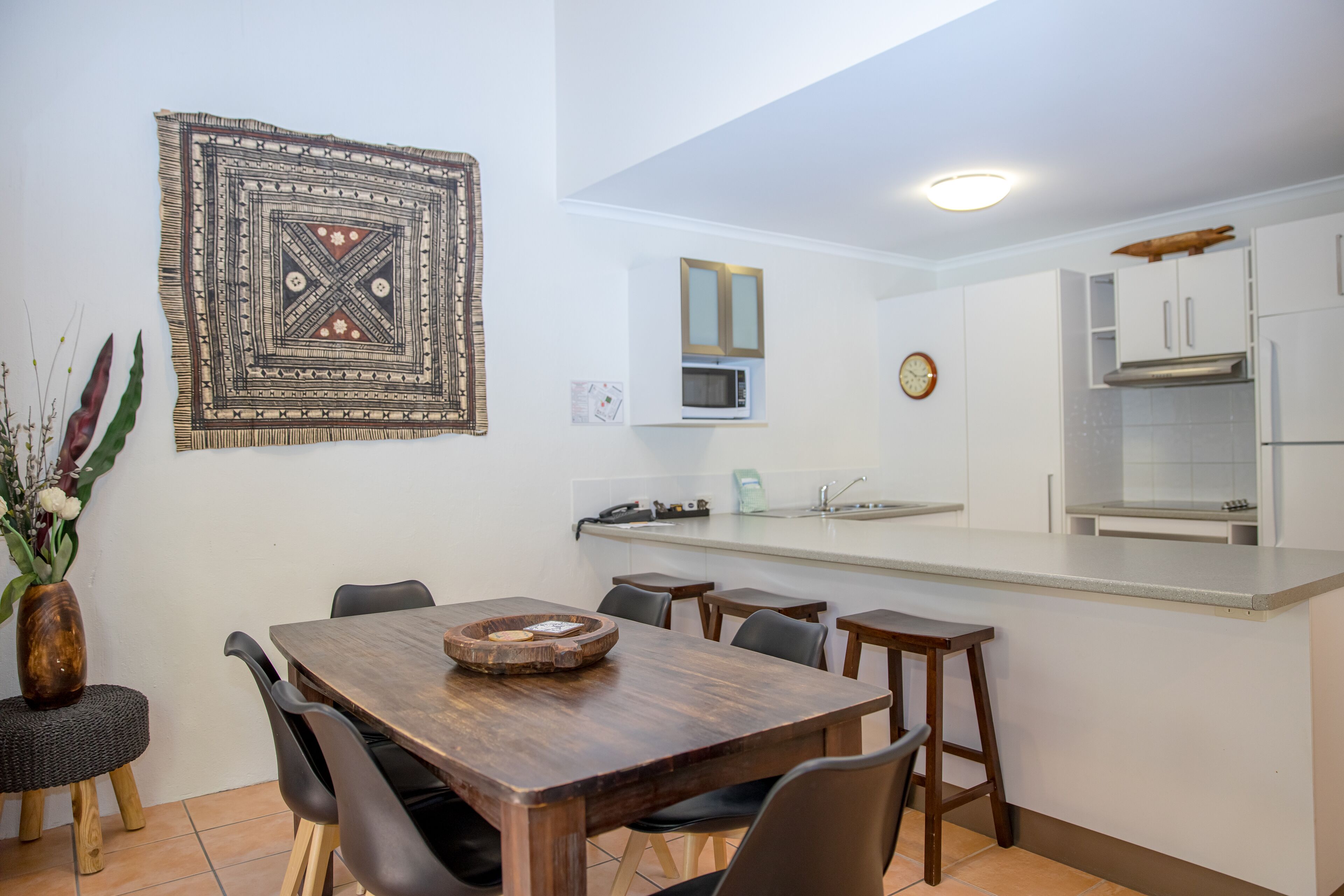 Une cuisine moderne avec des armoires blanches, un four à micro-ondes et une hotte de cuisine en acier inoxydable. On y trouve un coin repas avec une table et des chaises en bois, ainsi qu’une décoration murale.