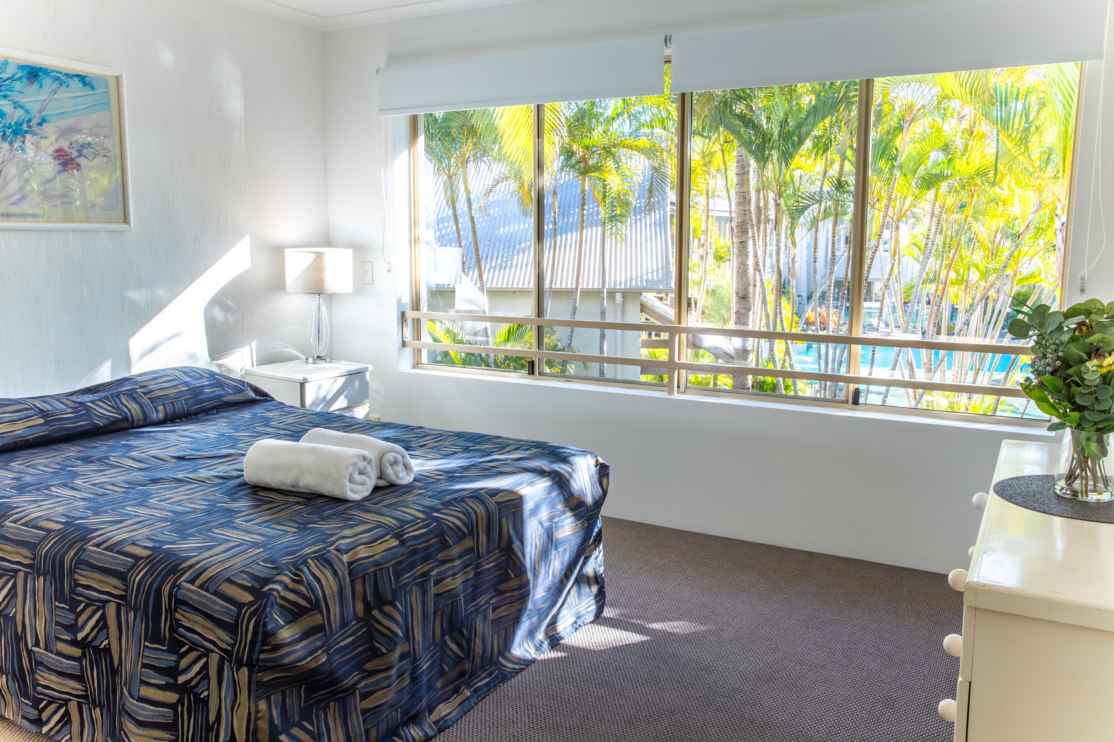 Habitación de hotel con una cama grande, una mesita de noche, una lámpara y vistas a palmeras y una piscina.