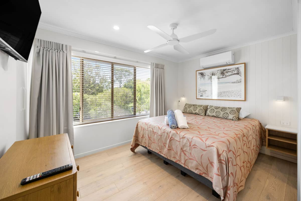 A bedroom with a bed, a ceiling fan, a window with blinds, a TV, and a remote control on a wooden table.
