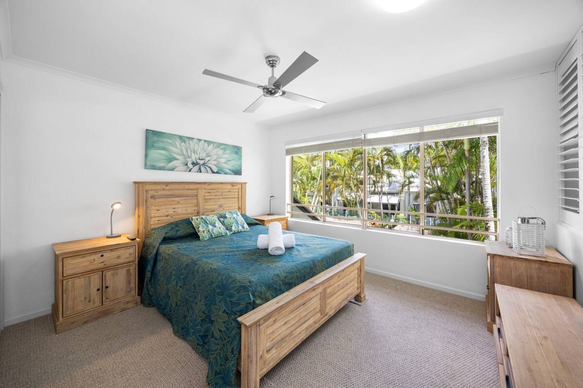 A bedroom with a bed, wooden headboard, bedside tables, a ceiling fan, a window with a view of palm trees, and a painting on the wall.