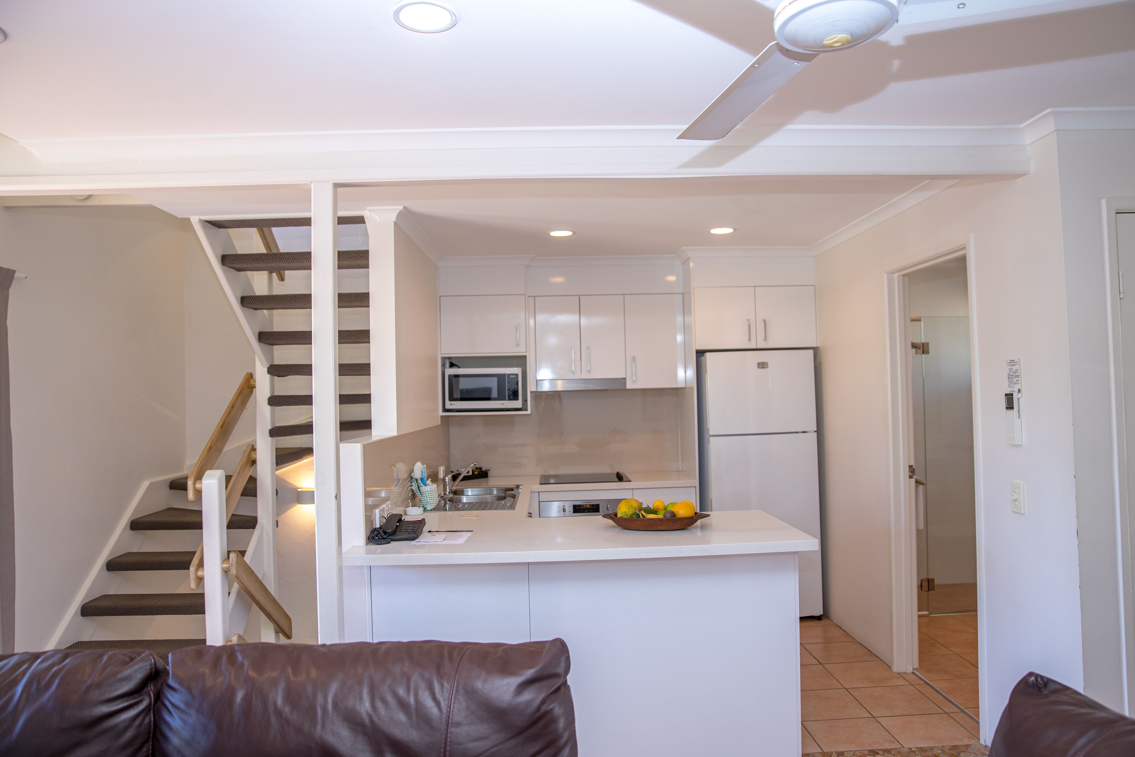Eine moderne Küche mit weißen Schränken, einer Mikrowelle und einem Kühlschrank. In der Mitte steht eine kleine Kochinsel mit einer Obstschale. Eine Treppe mit Holzsitzstufen und weißen Geländern ist sichtbar.