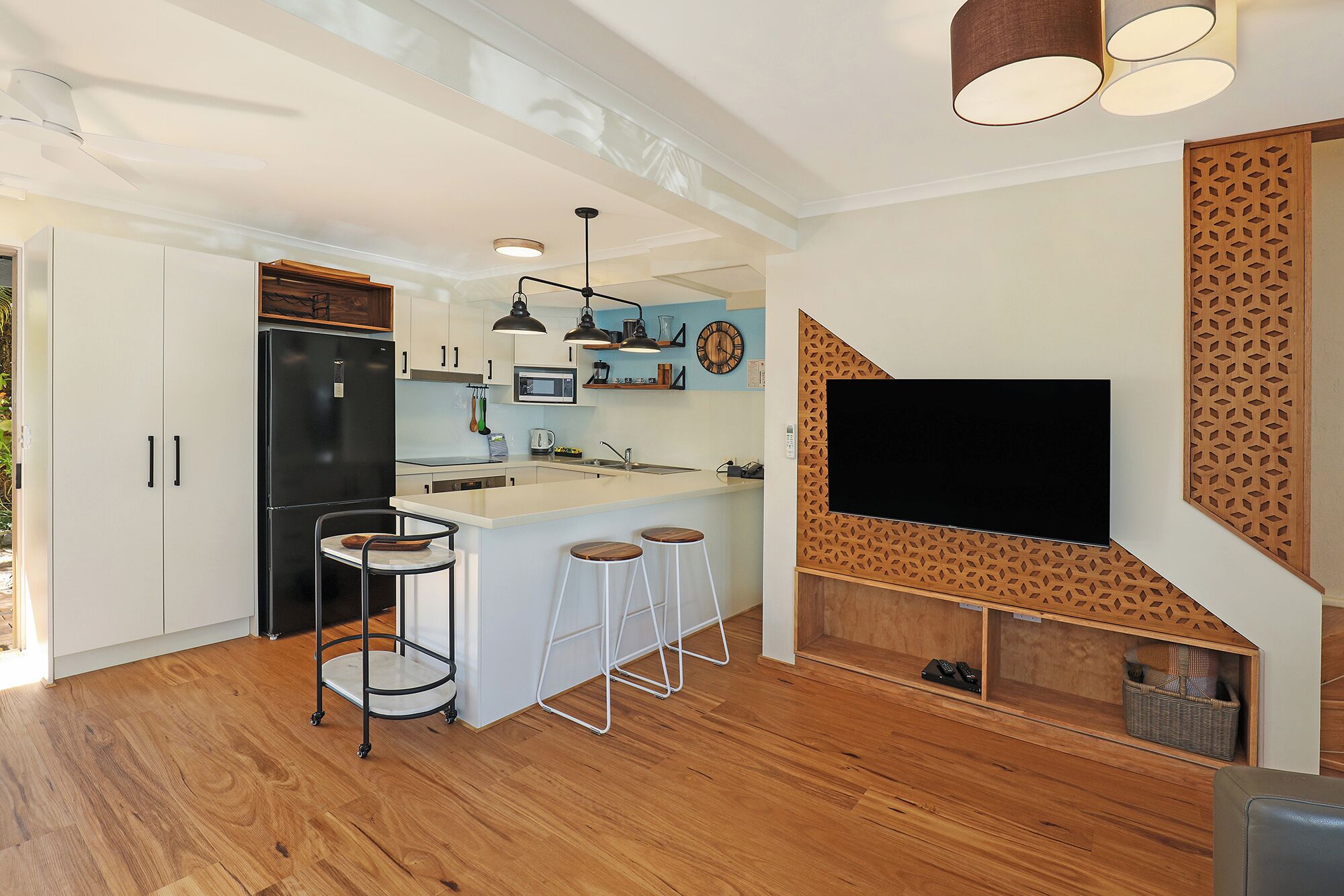 A modern kitchen with white cabinets, a black refrigerator, a central island with stools, pendant lighting, and a large flat-screen TV mounted on a wooden wall with a geometric pattern.