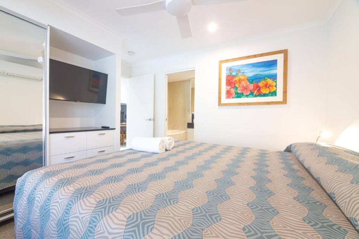 A hotel room with a bed, a TV, a painting, and a ceiling fan.