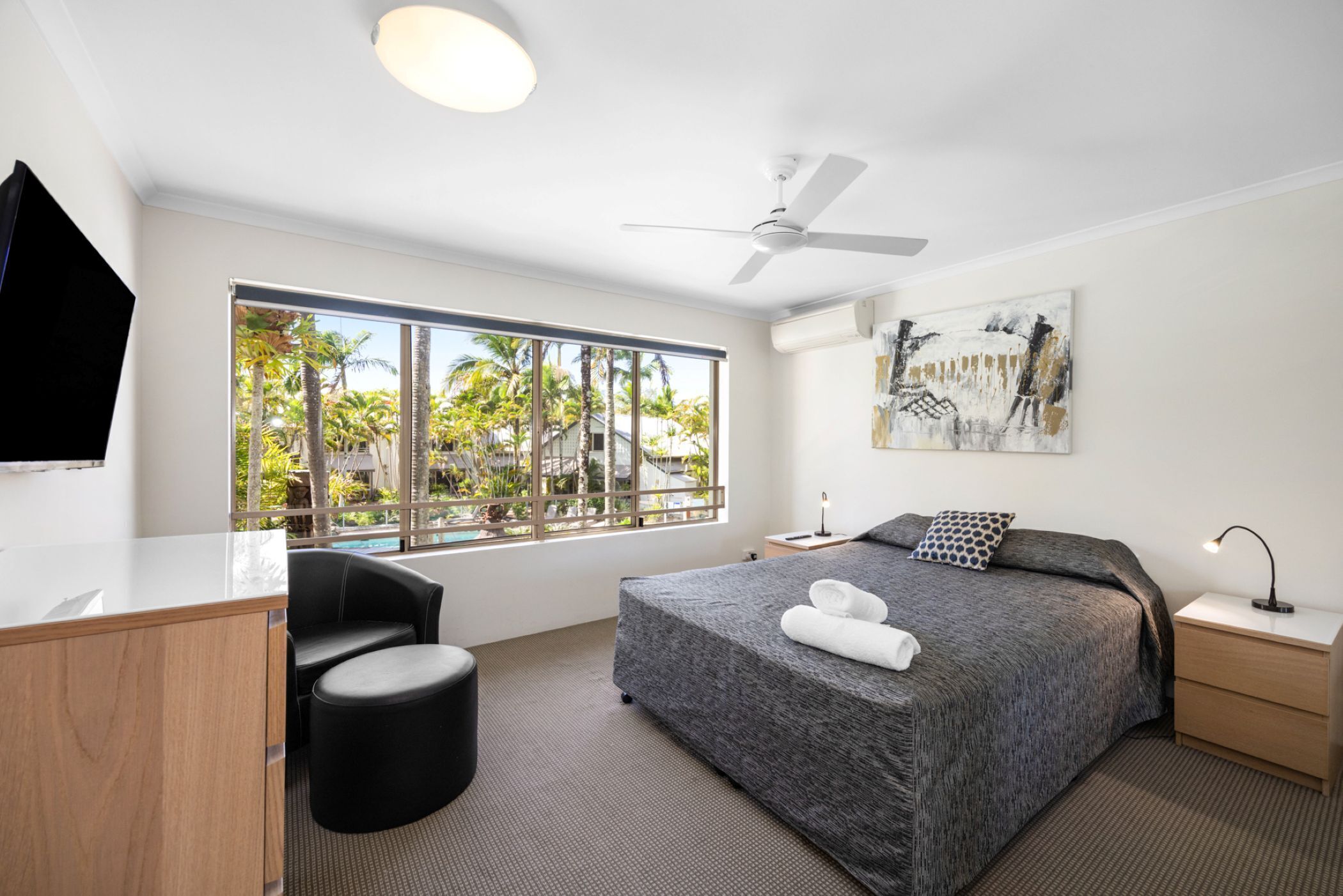 A modern hotel room with a large bed, a desk, a chair, a ceiling fan, and a large window with a view of palm trees.