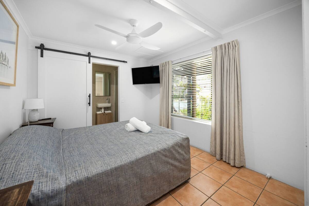 A bedroom with a bed, a ceiling fan, a TV, a window with curtains, and a door.