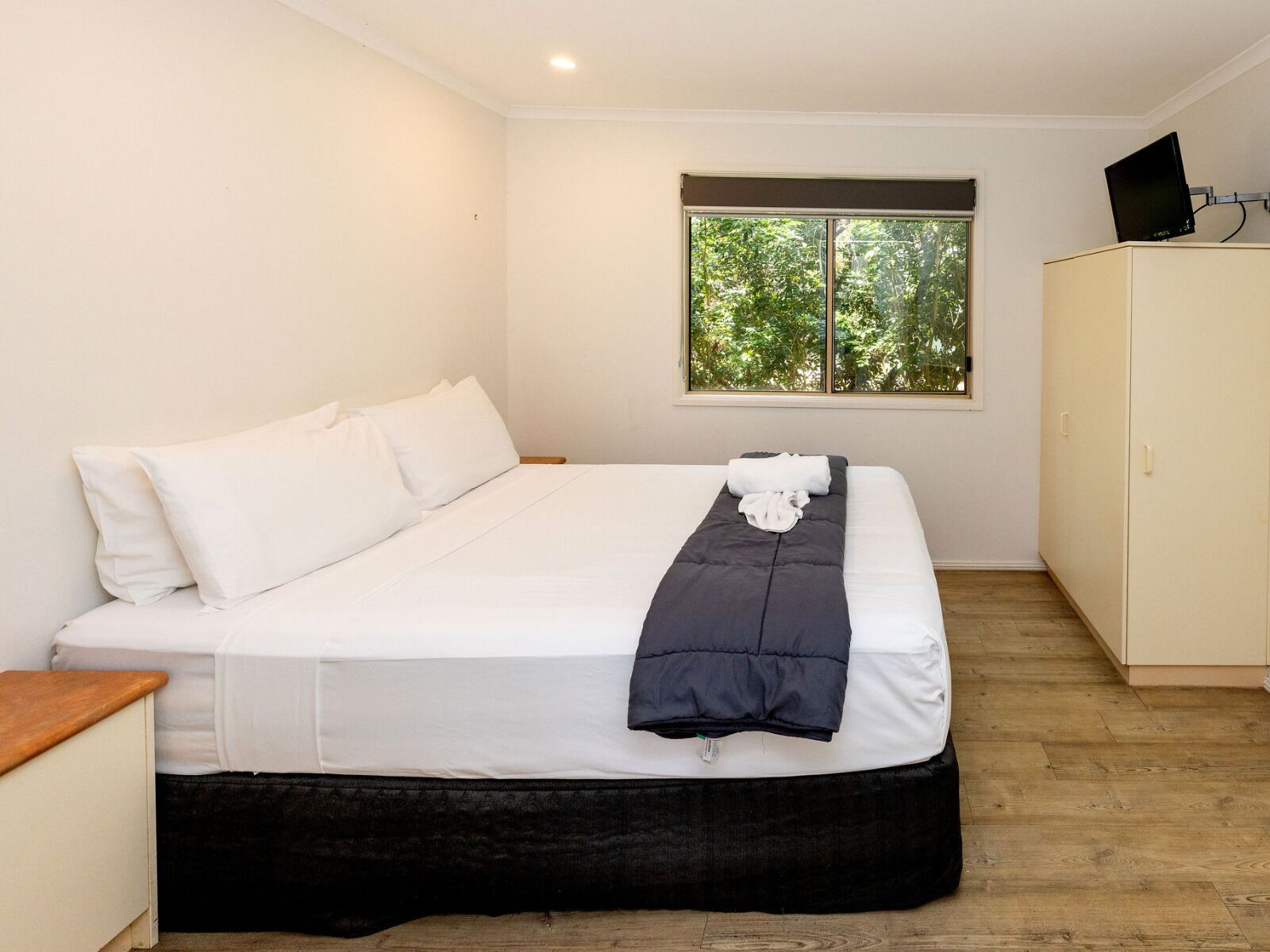Una cama bien hecha con ropa de cama blanca y oscura, una ventana con vistas a árboles, y un televisor sobre un mueble.