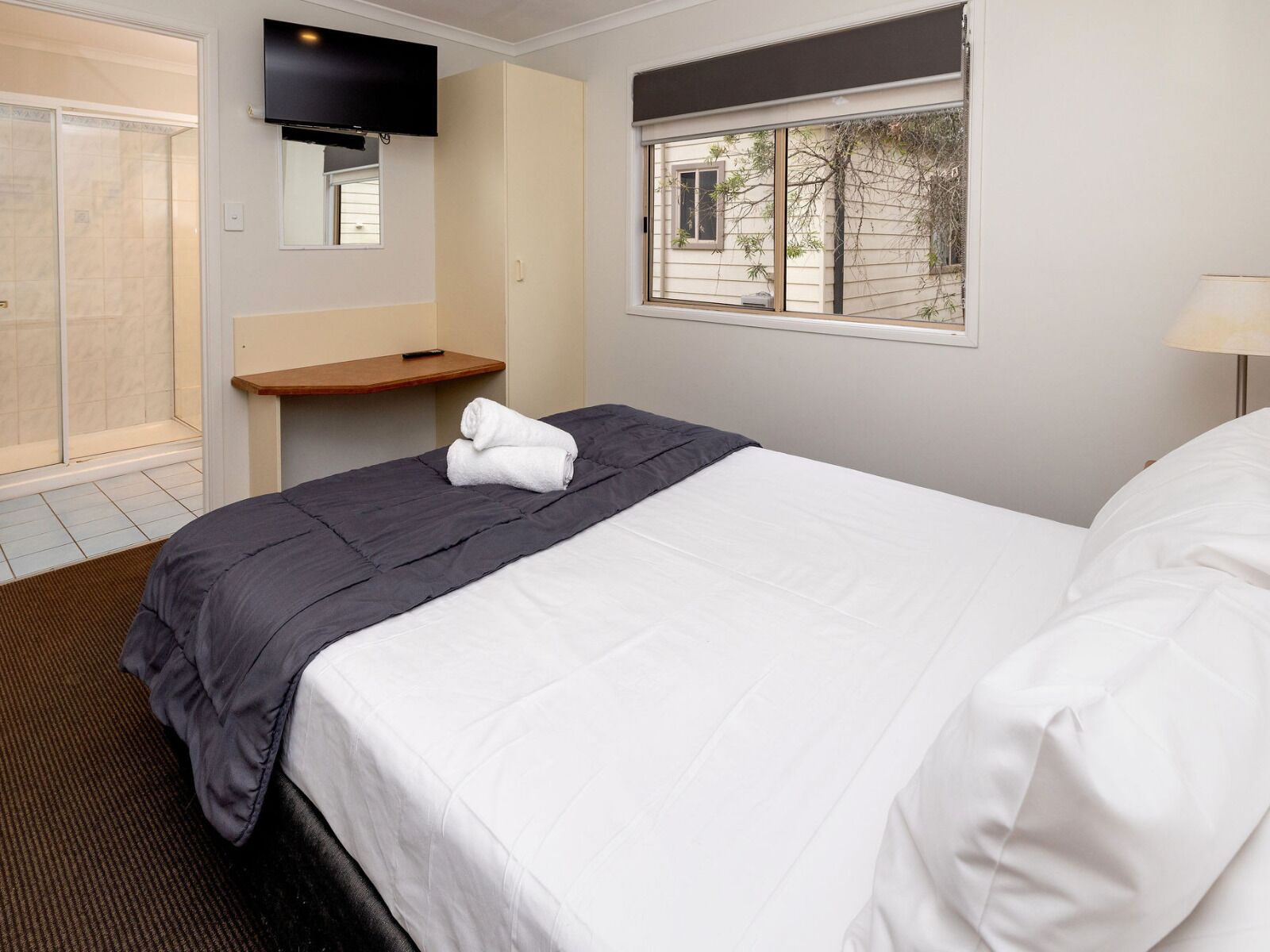 A hotel room with a bed, a bedside table, a TV mounted on the wall, a window with a view of the outside, and a shower area.