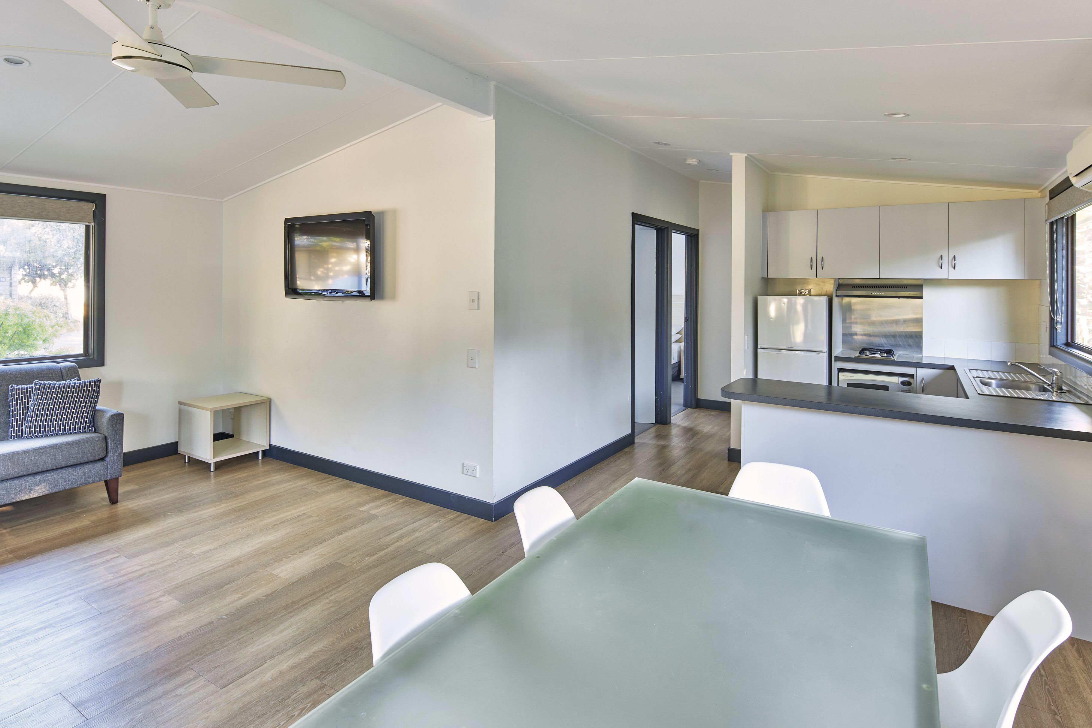 A modern kitchen with white cabinets, a stainless steel refrigerator, and a built-in oven. There is a dining area with a table and chairs, and a ceiling fan.