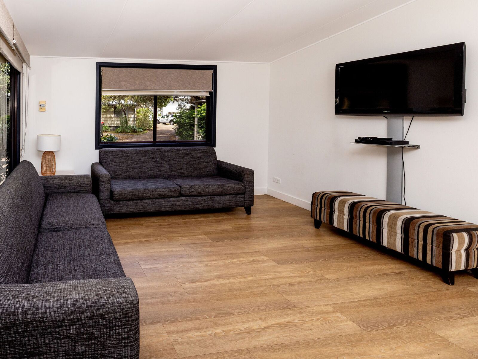 A modern living room with a grey sofa, a striped bench, a flat-screen TV mounted on the wall, and a large window with a view of the outdoors.