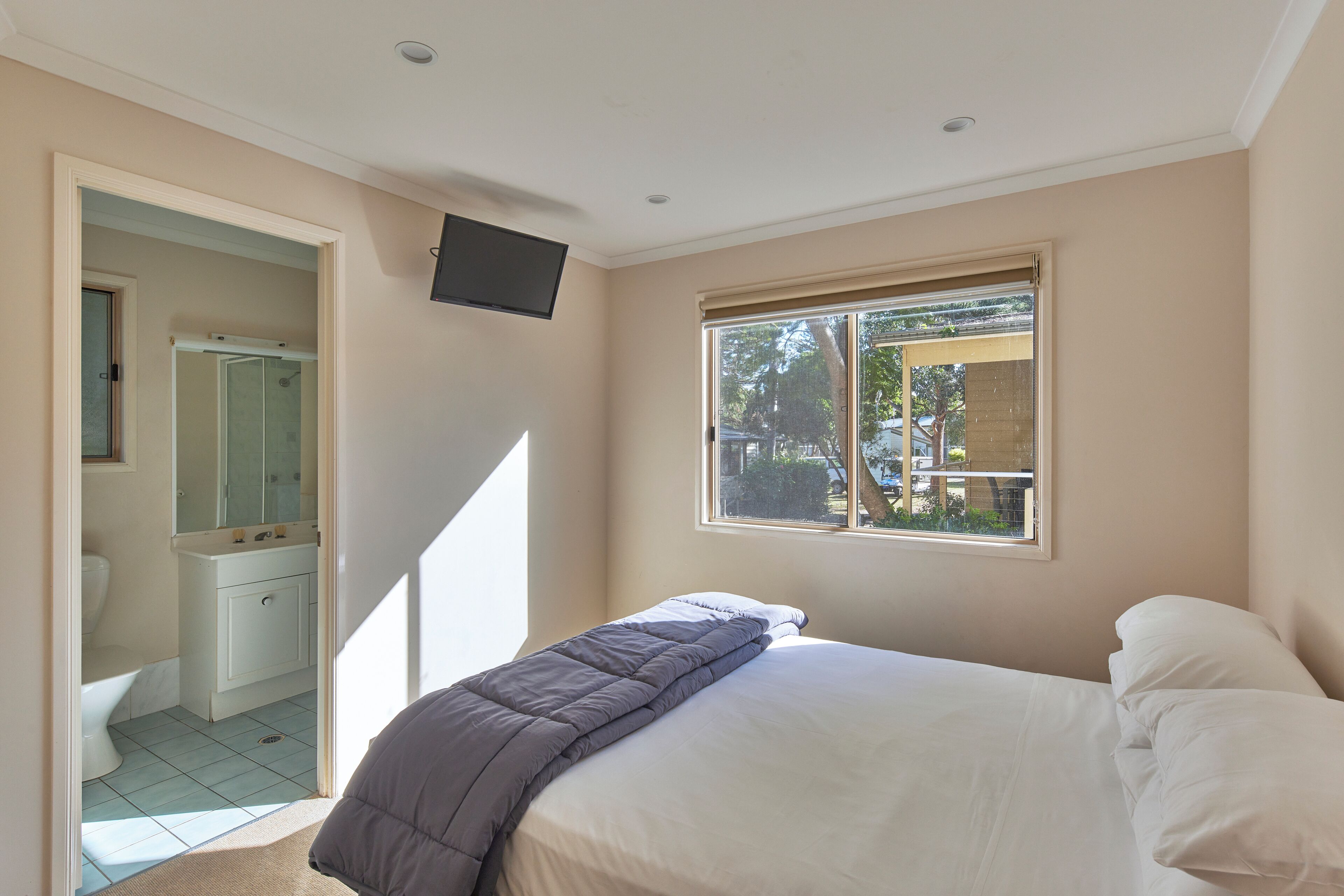 Un dormitorio con cama, ventana, televisor montado en la pared y baño visible a través de una puerta abierta.