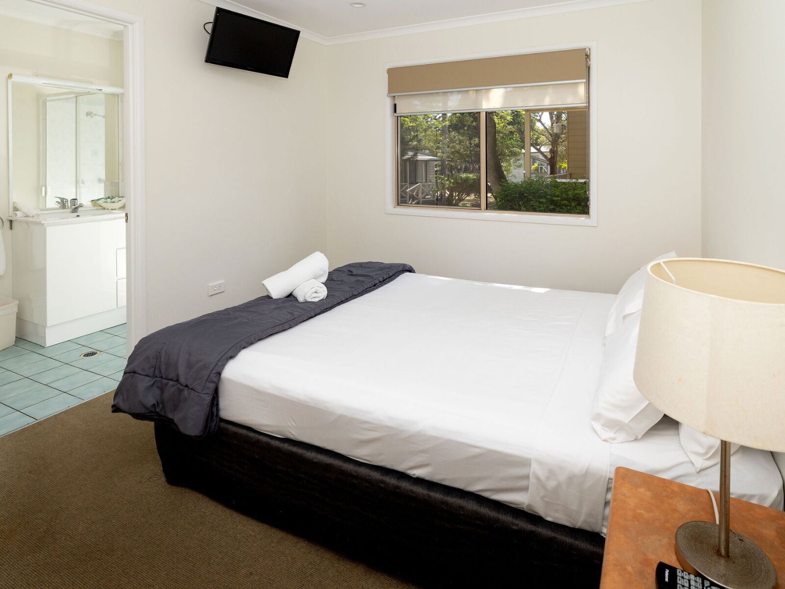 A hotel room with a bed, a TV mounted on the wall, a window with a view of trees, and a bathroom visible through an open door.