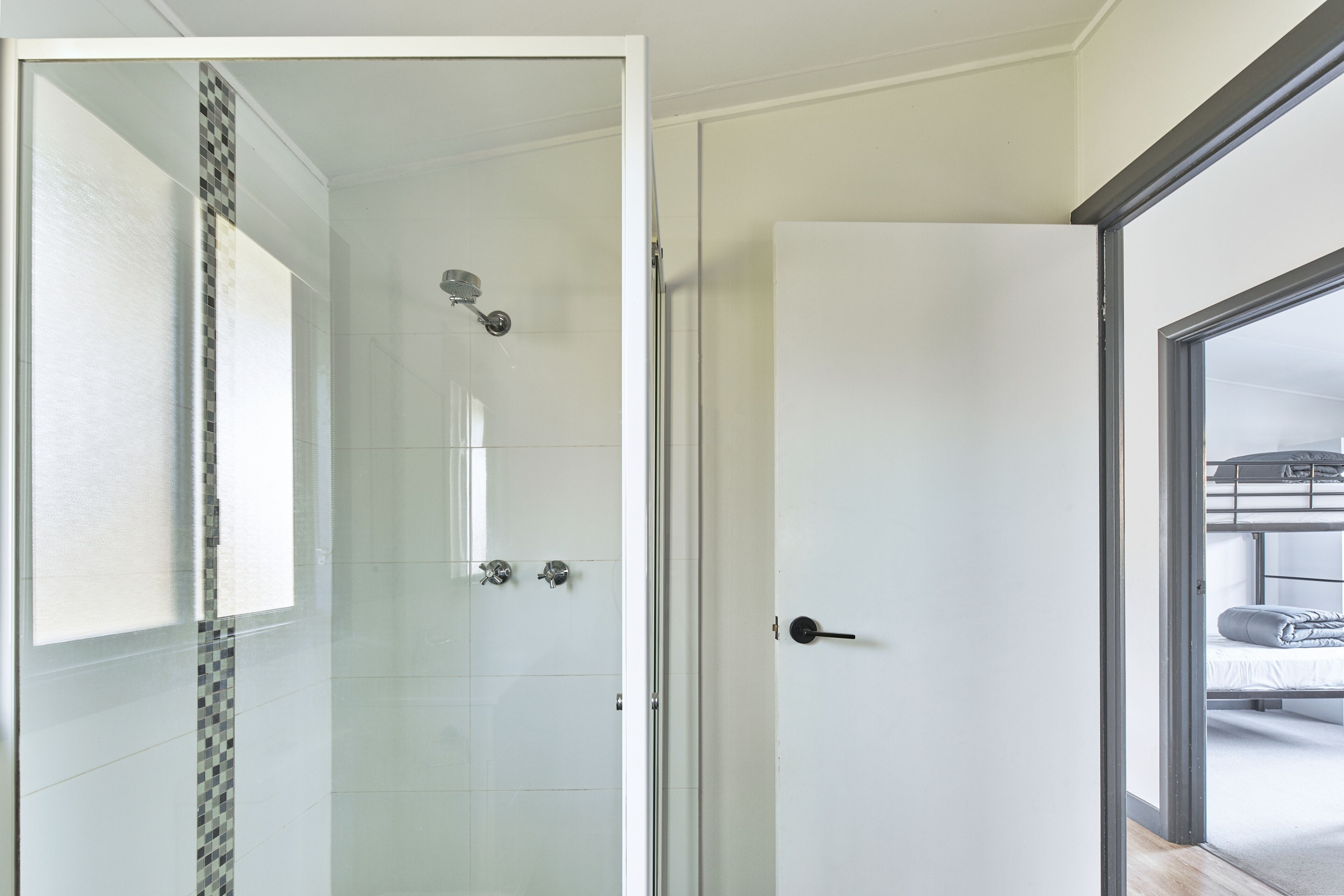A modern bathroom with a glass shower enclosure, a white door, and a view of a bunk bed room through an open door.