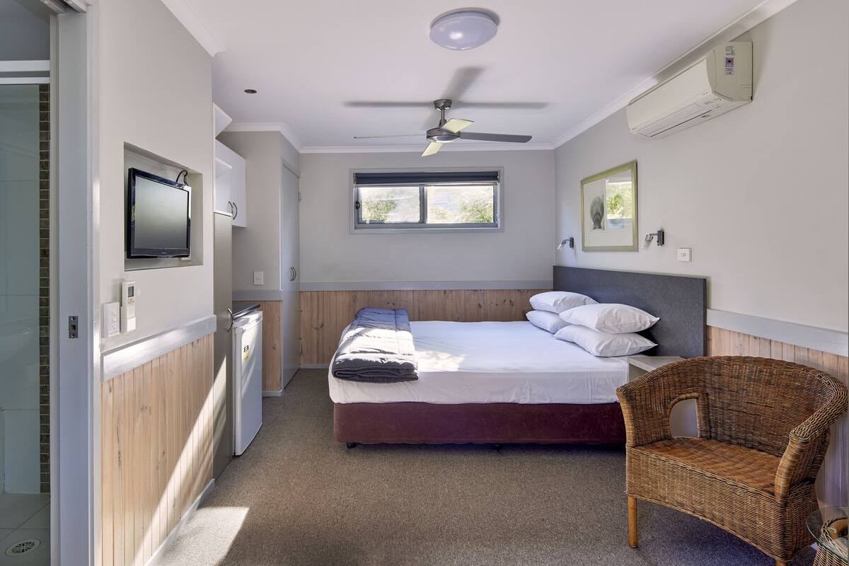 Een slaapkamer met een bed, een stoel, een televisie, een plafondventilator, een raam en een airconditioner.