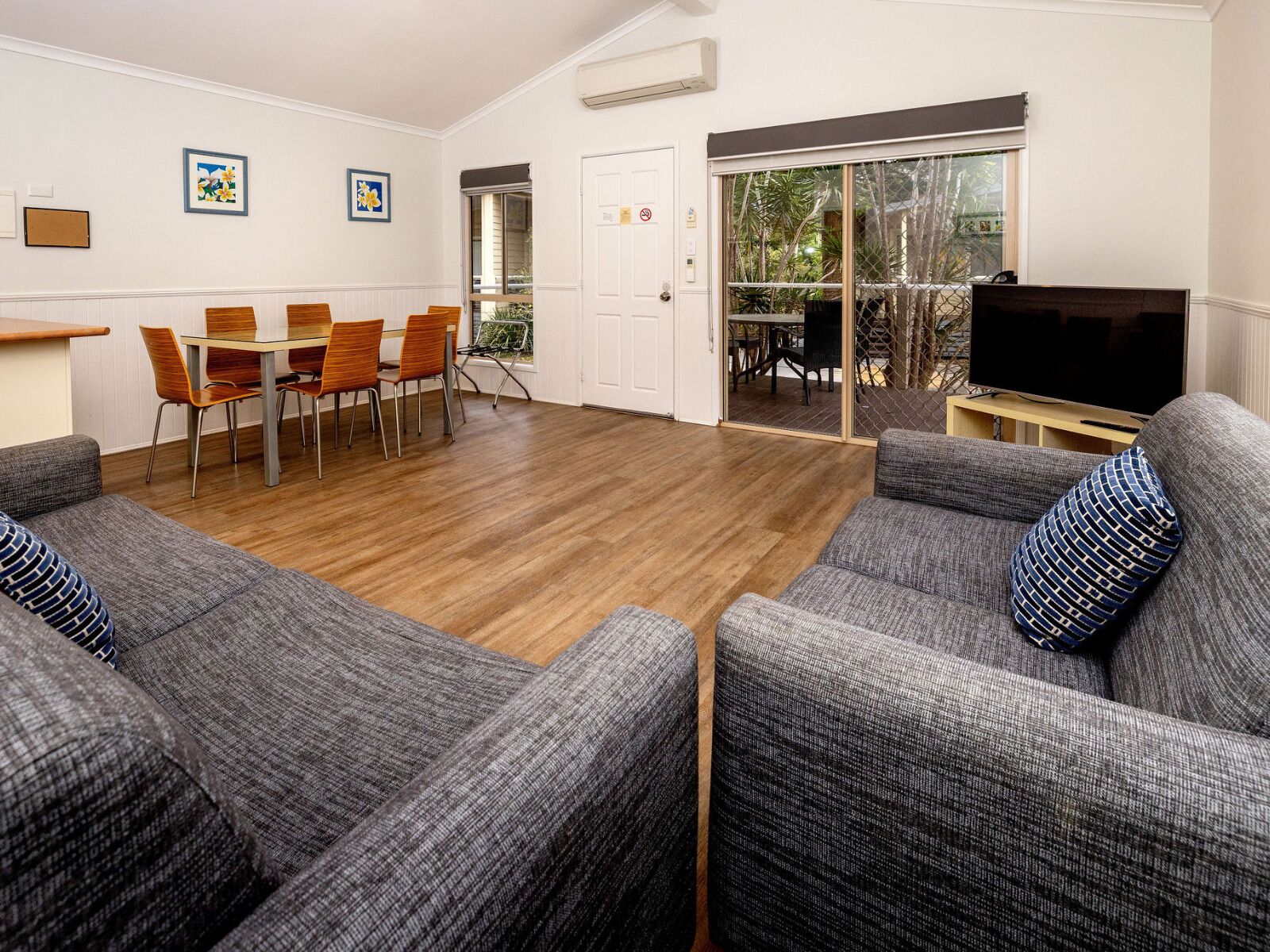 A modern living room with grey upholstered sofas, a wooden dining table with chairs, a flat-screen TV, and a sliding glass door leading to an outdoor area.