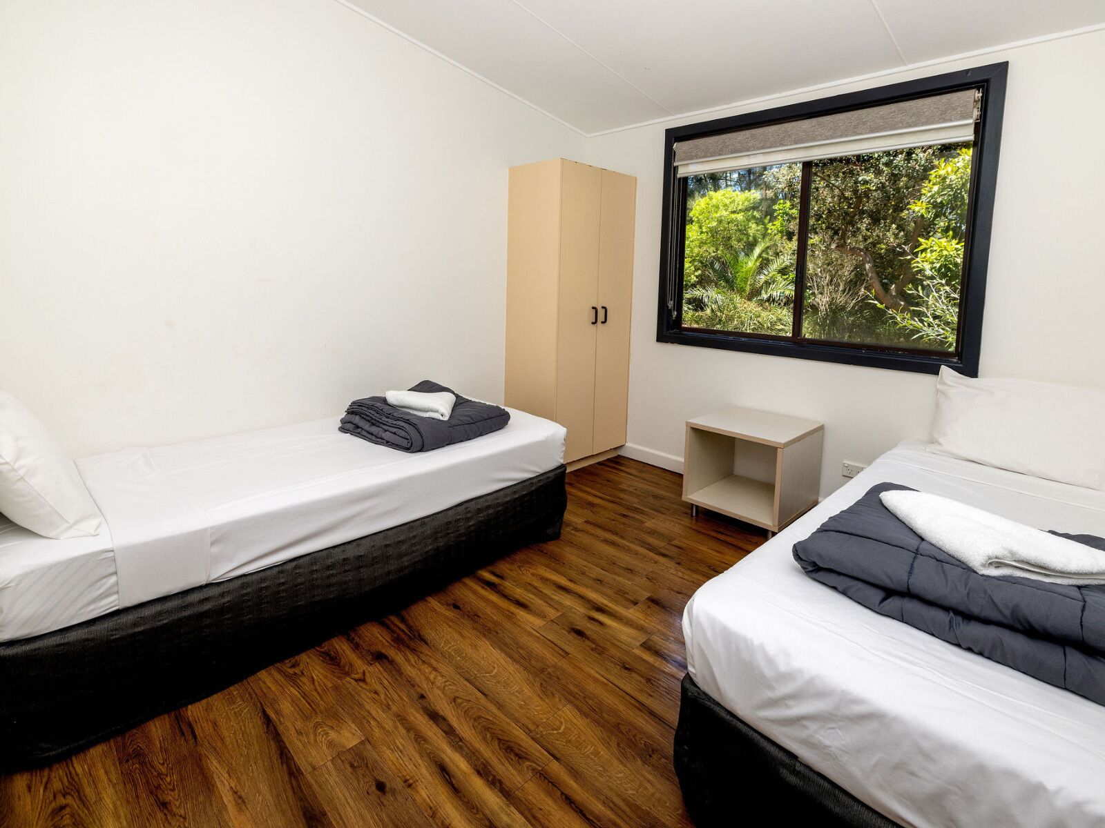 A bedroom with two beds, a wardrobe, a small table, and a window with a view of greenery.