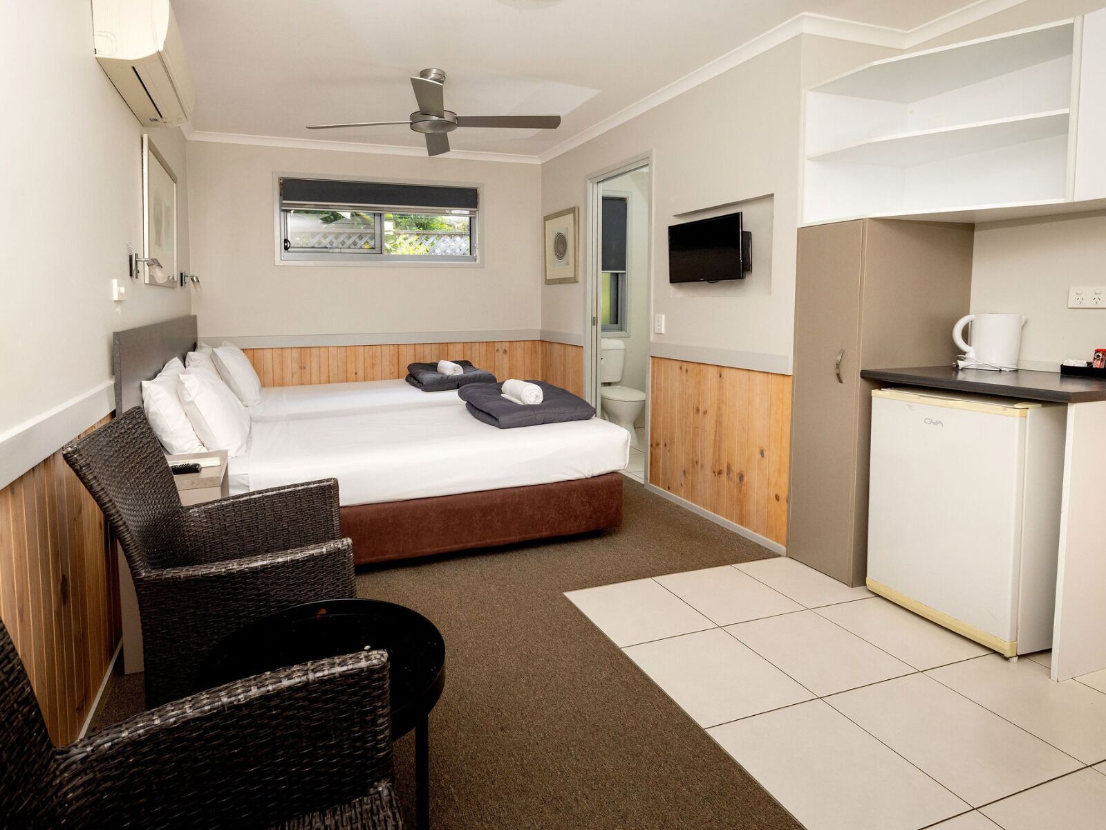 A hotel room with a bed, a desk, a chair, a kitchenette, and a ceiling fan.
