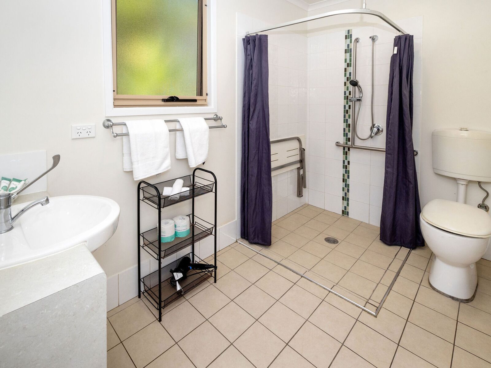 Un baño con lavamanos, un estante con artículos de aseo, ducha con barra de apoyo y un inodoro.