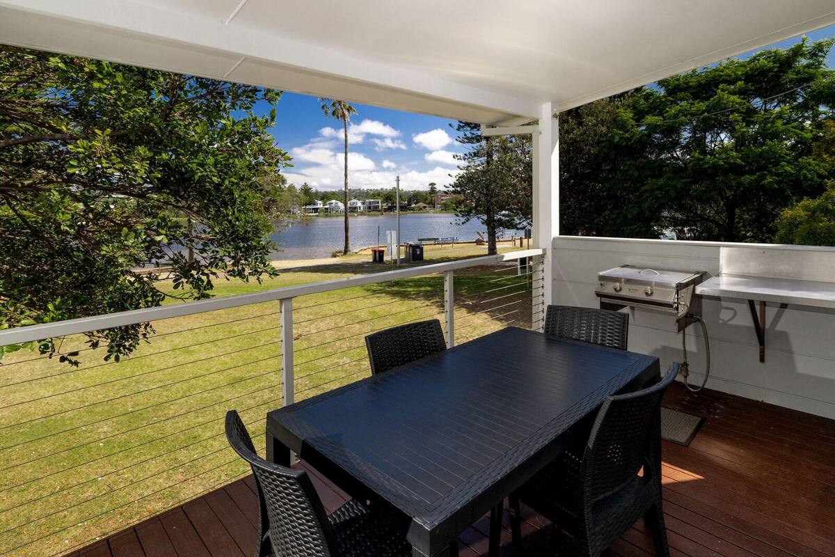 Een overdekt terras buiten met een tafel en stoelen, met uitzicht op een meer en een gazon.