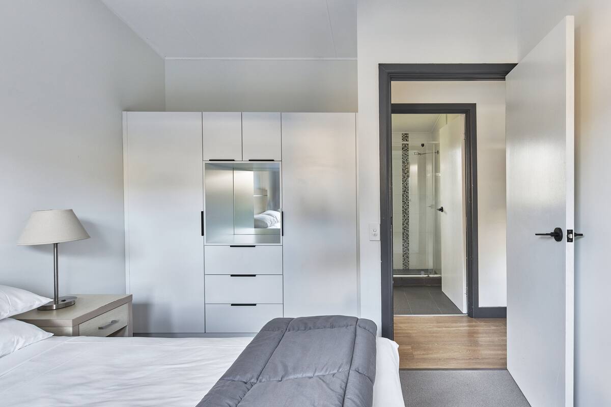 Een moderne slaapkamer met een bed, nachtkastje, lamp, garderobe, en een zichtbare badkamer door een geopende deur.