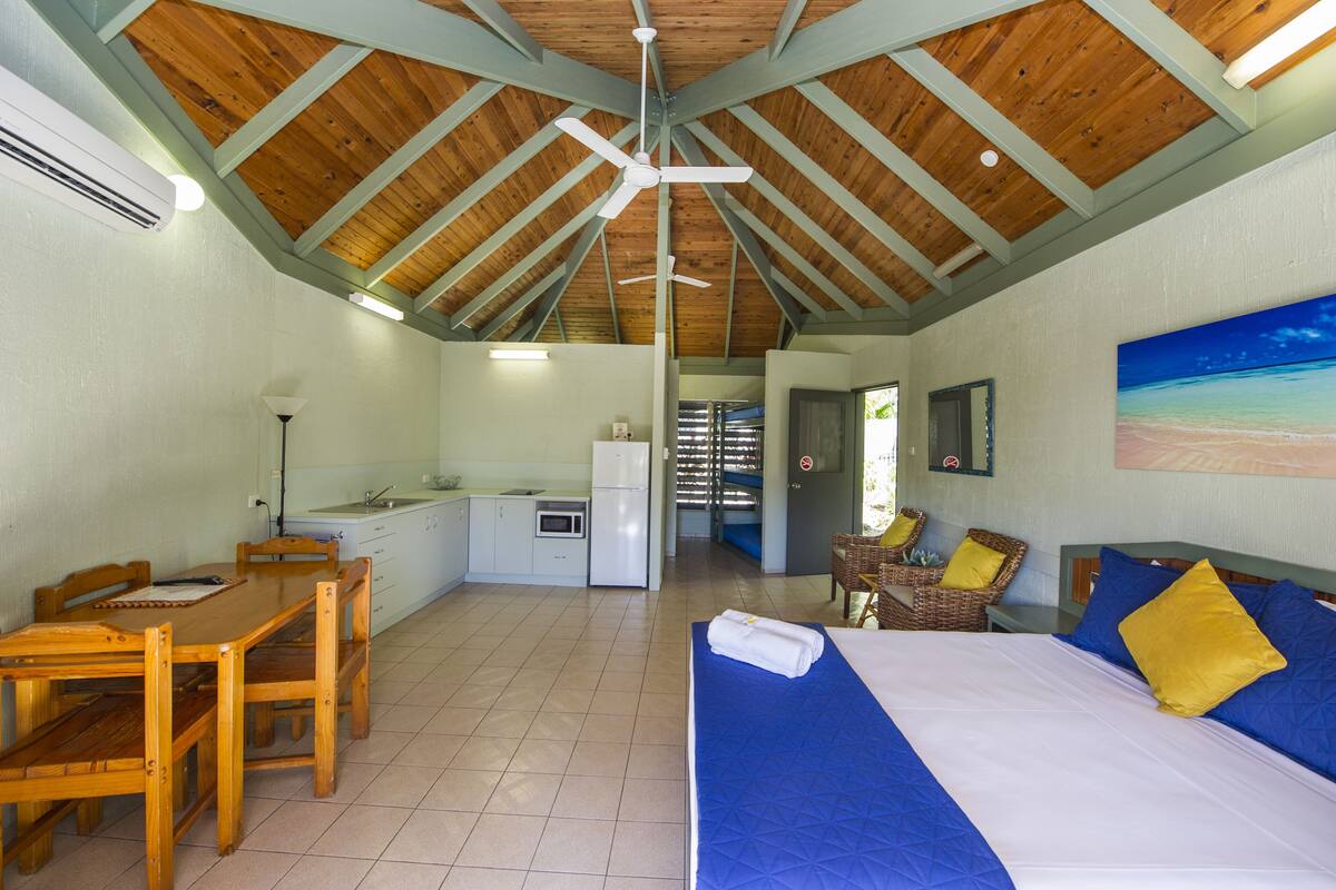 A room with a wooden ceiling, a bed with blue and yellow pillows, a dining table with chairs, a kitchenette, and a painting on the wall.
