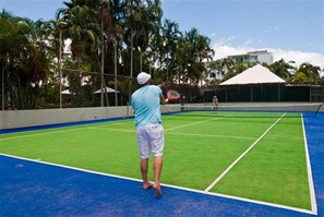 Una cancha de tenis con red, rodeada de palmeras y un edificio al fondo.