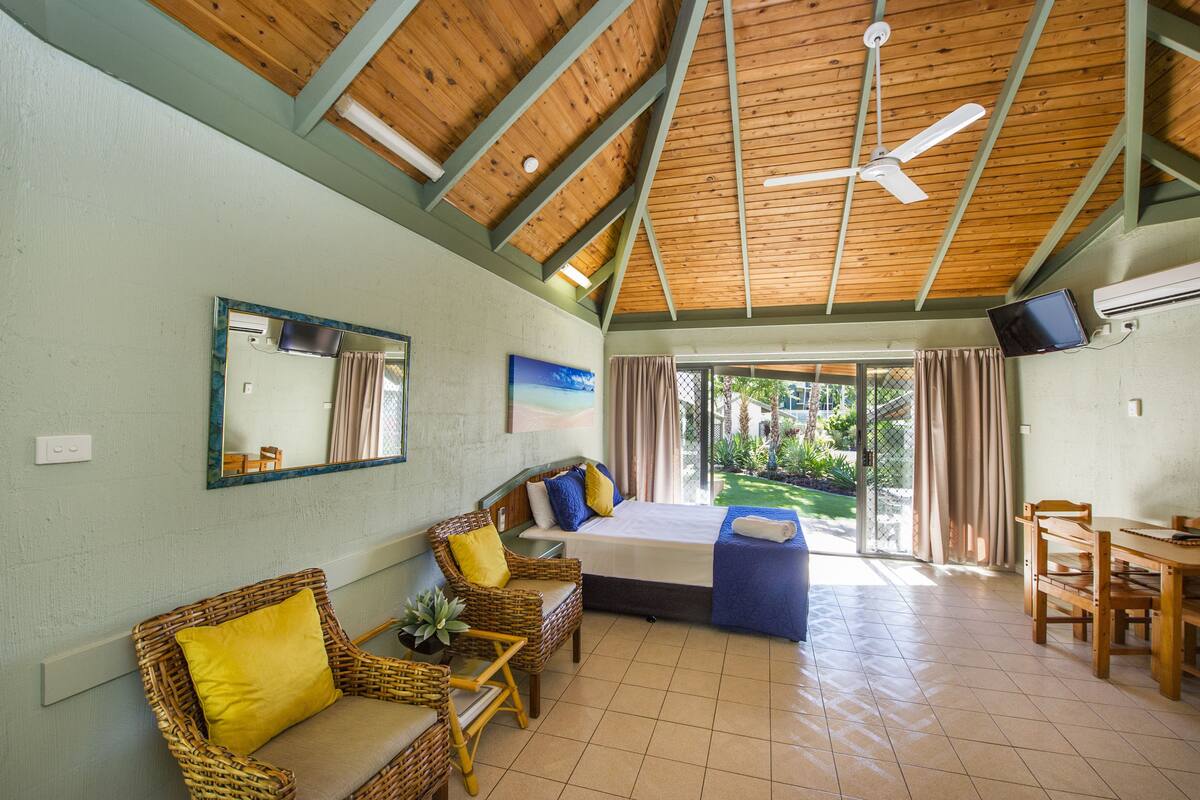 A room with a bed, two wicker chairs with cushions, a small table, and a ceiling fan.