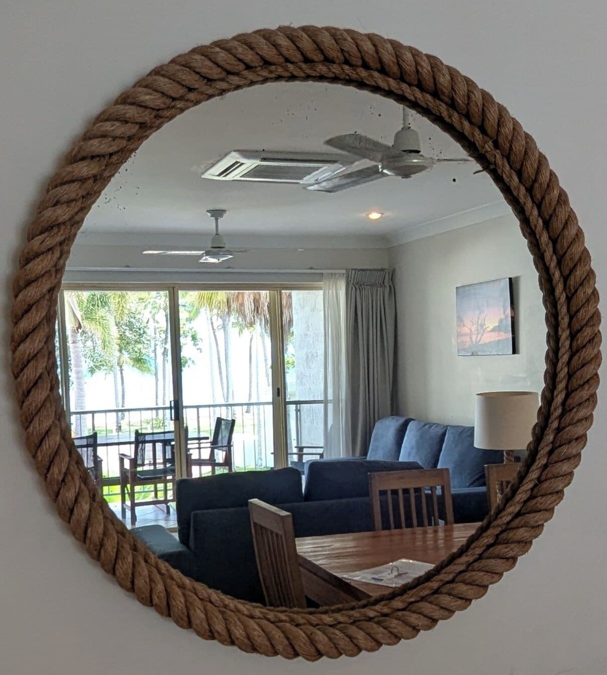 A room with a round mirror, a ceiling fan, a sofa, a table, and a view of palm trees outside.