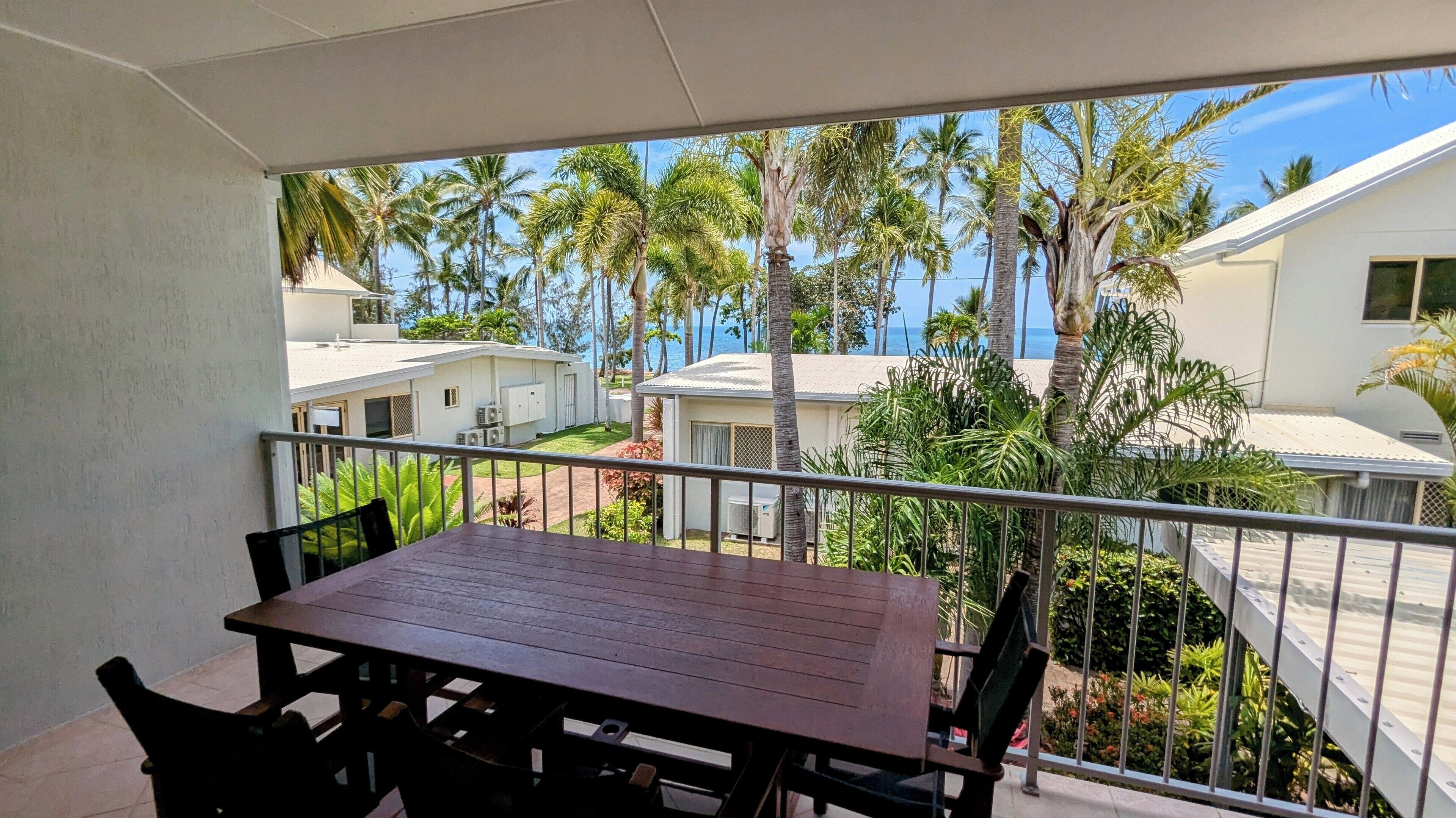 A balcony with a wooden table and chairs, overlooking a residential area with palm trees and houses.