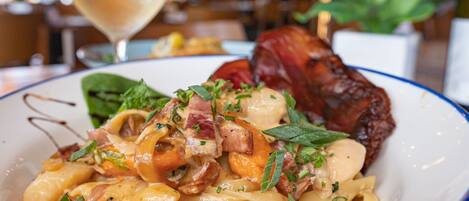 Une assiette de pâtes avec des lardons et des légumes verts, un verre de vin blanc et une plante en arrière-plan.