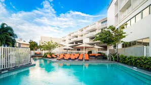 Una piscina con sillas de descanso anaranjadas, una cerca blanca y un edificio de varios pisos con balcones.