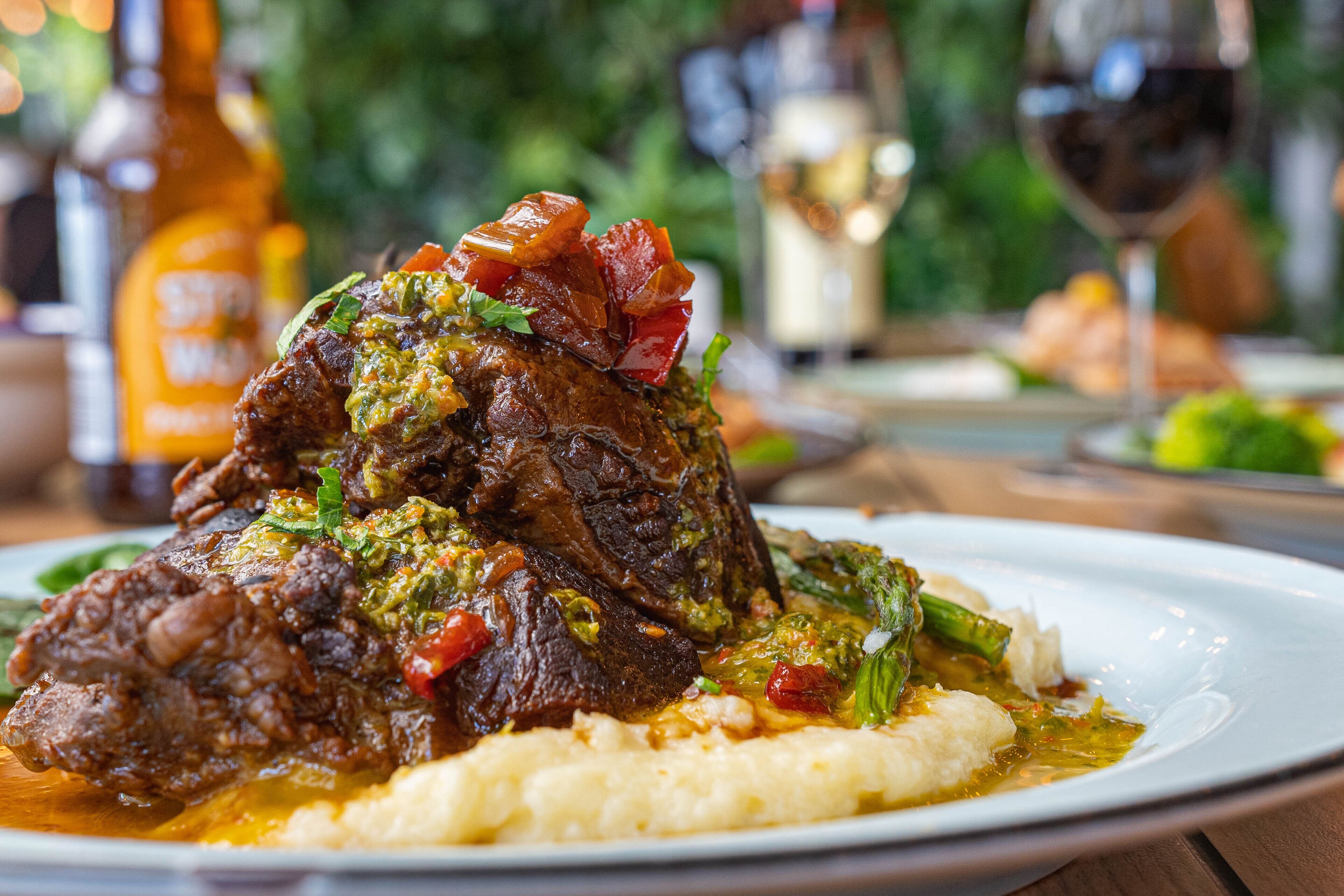 Une assiette de nourriture comprenant un plat de viande, des légumes et un accompagnement crémeux, posée sur une table avec une bouteille et un verre en arrière-plan.