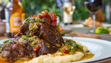 Une assiette de nourriture comprenant un plat de viande, des légumes et un accompagnement crémeux, posée sur une table avec une bouteille et un verre en arrière-plan.