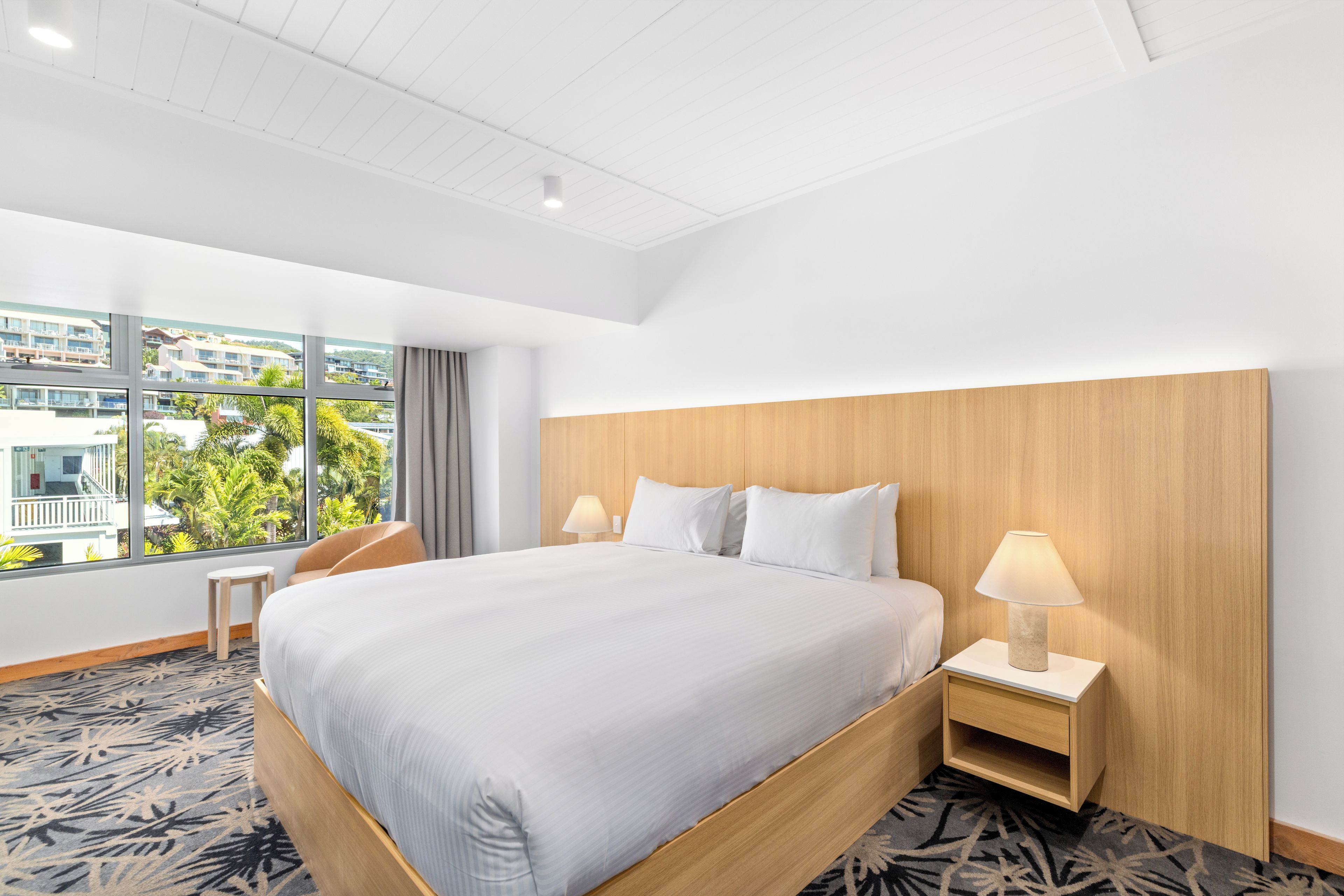 Une chambre d’hôtel avec un grand lit, deux tables de chevet, une vue sur un immeuble par la fenêtre et un plafond orné d’un motif en réseau.