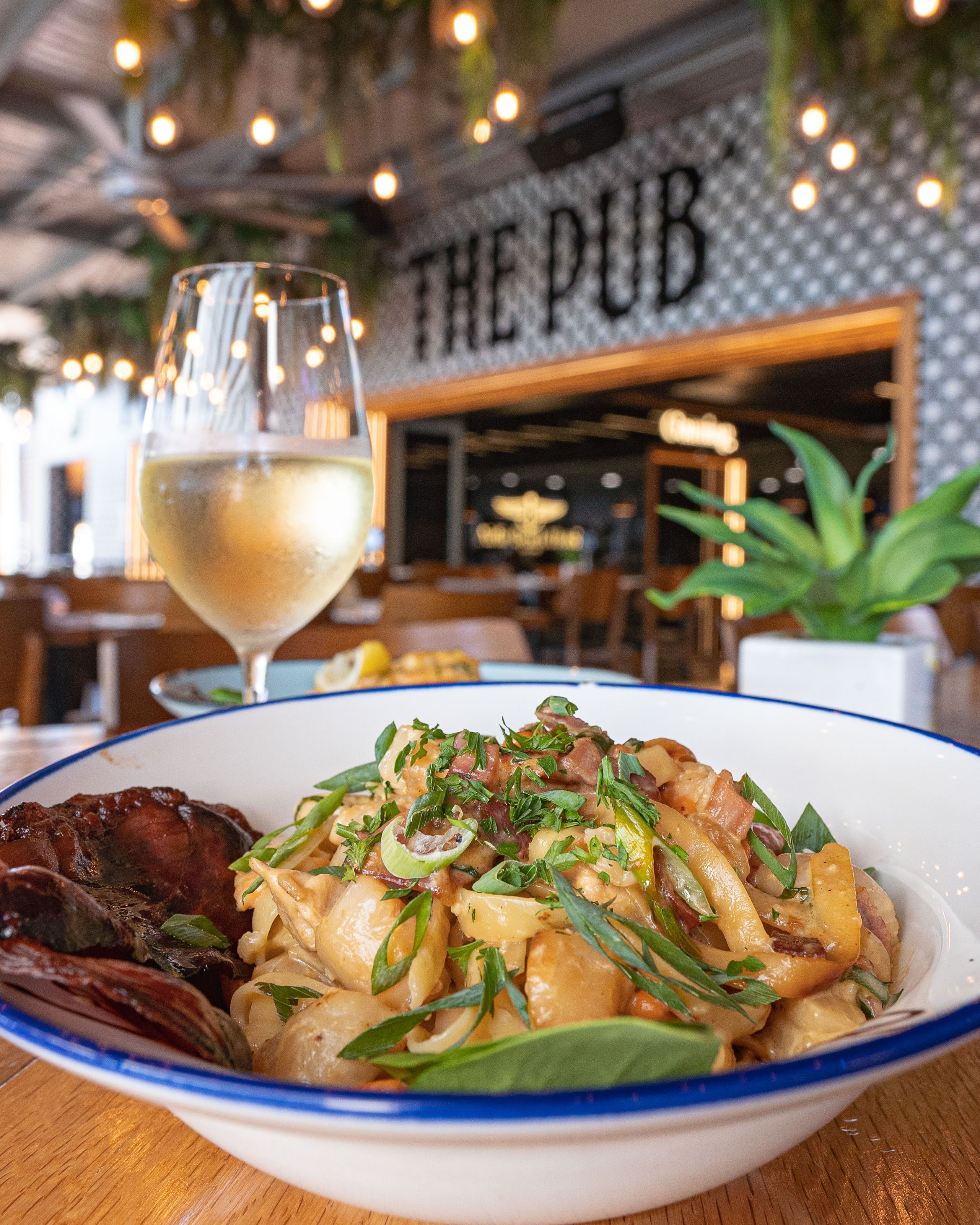 Uma tigela de massa com carne e vegetais, um copo de vinho branco, e um restaurante chamado "THE PUB" ao fundo.