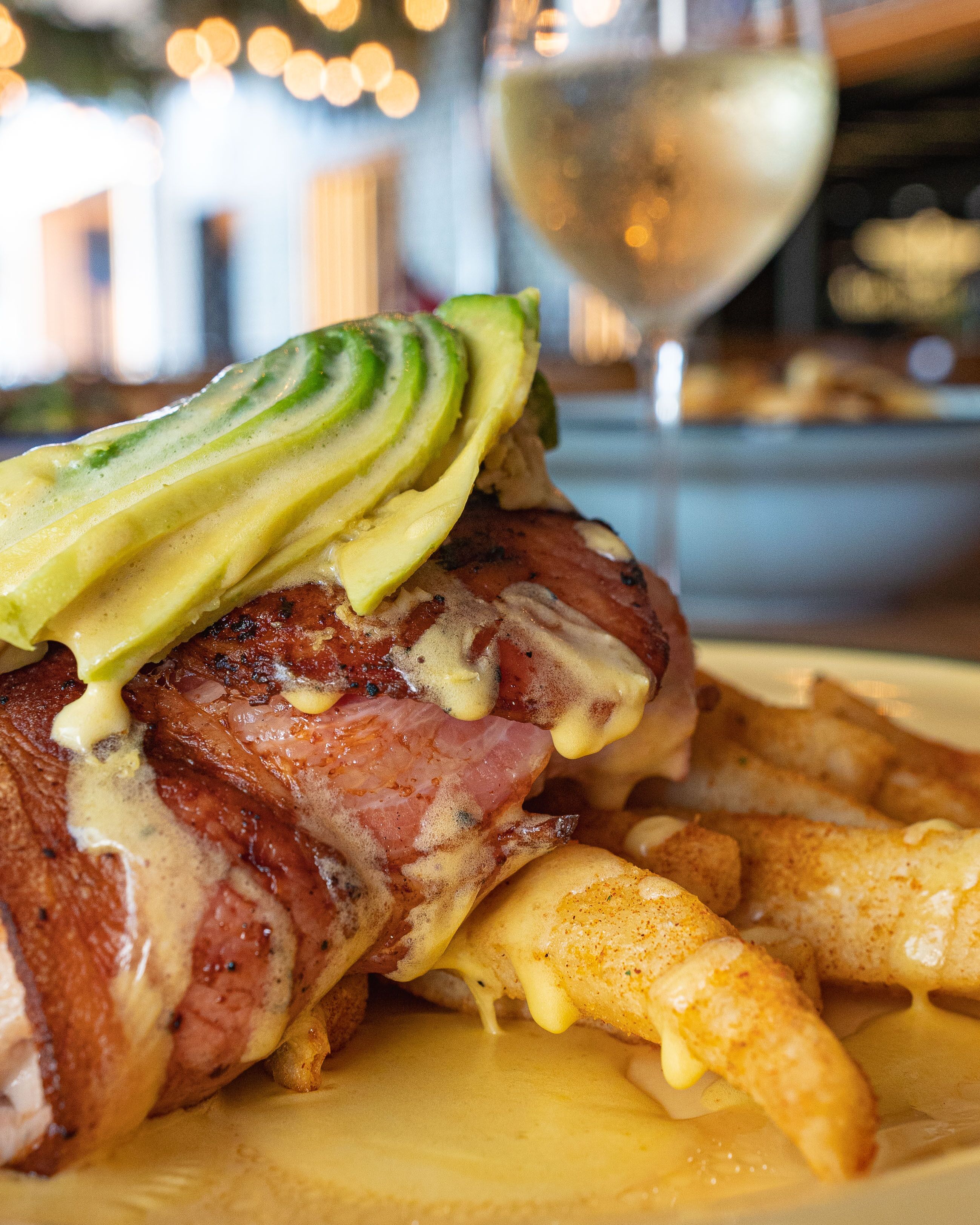 A plate with a steak topped with avocado slices and melted cheese, accompanied by fries and a glass of white wine.