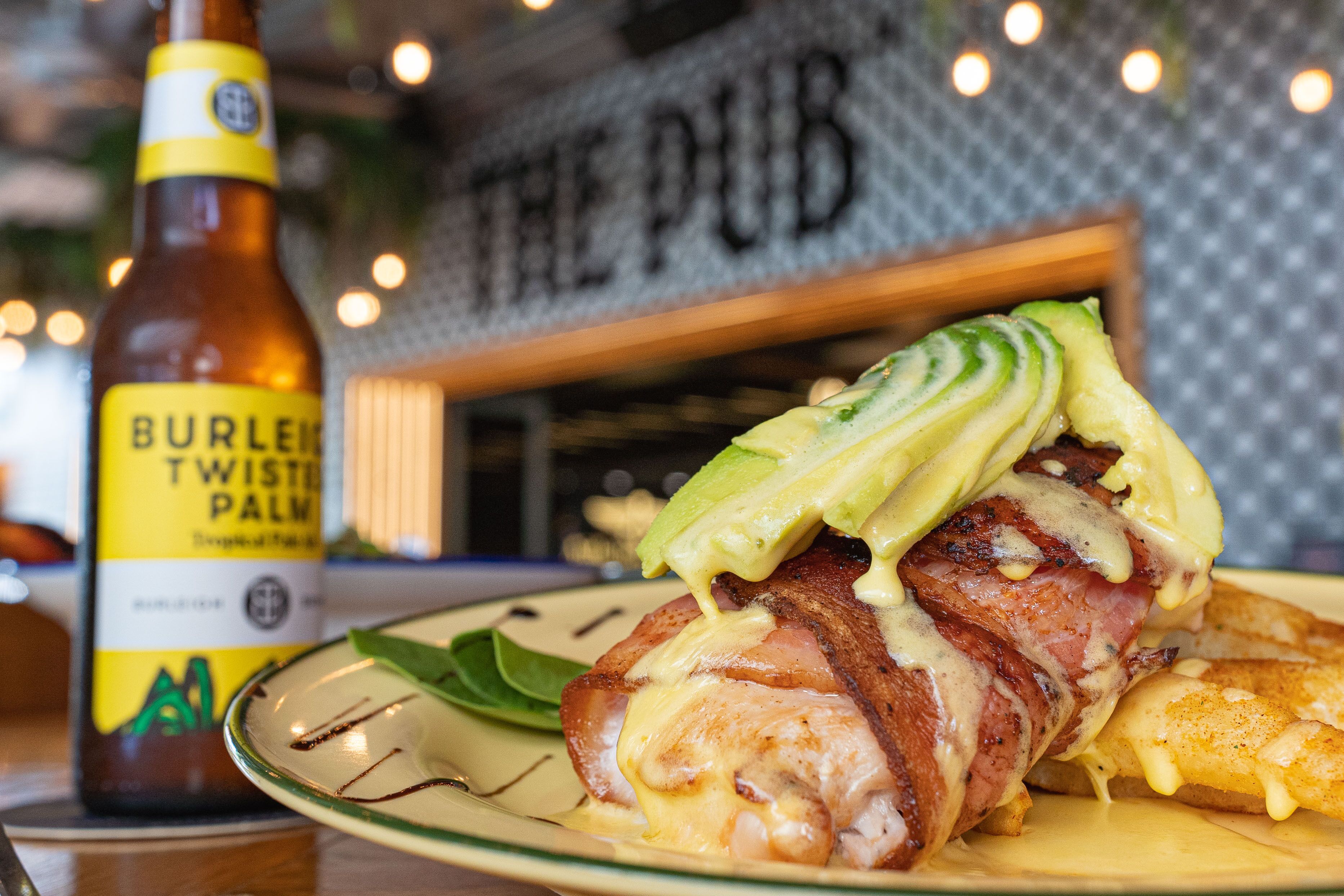 A plate with a wrapped chicken breast, avocado slices, and melted cheese. A bottle of Burley Twisted Palm beer is visible in the background.