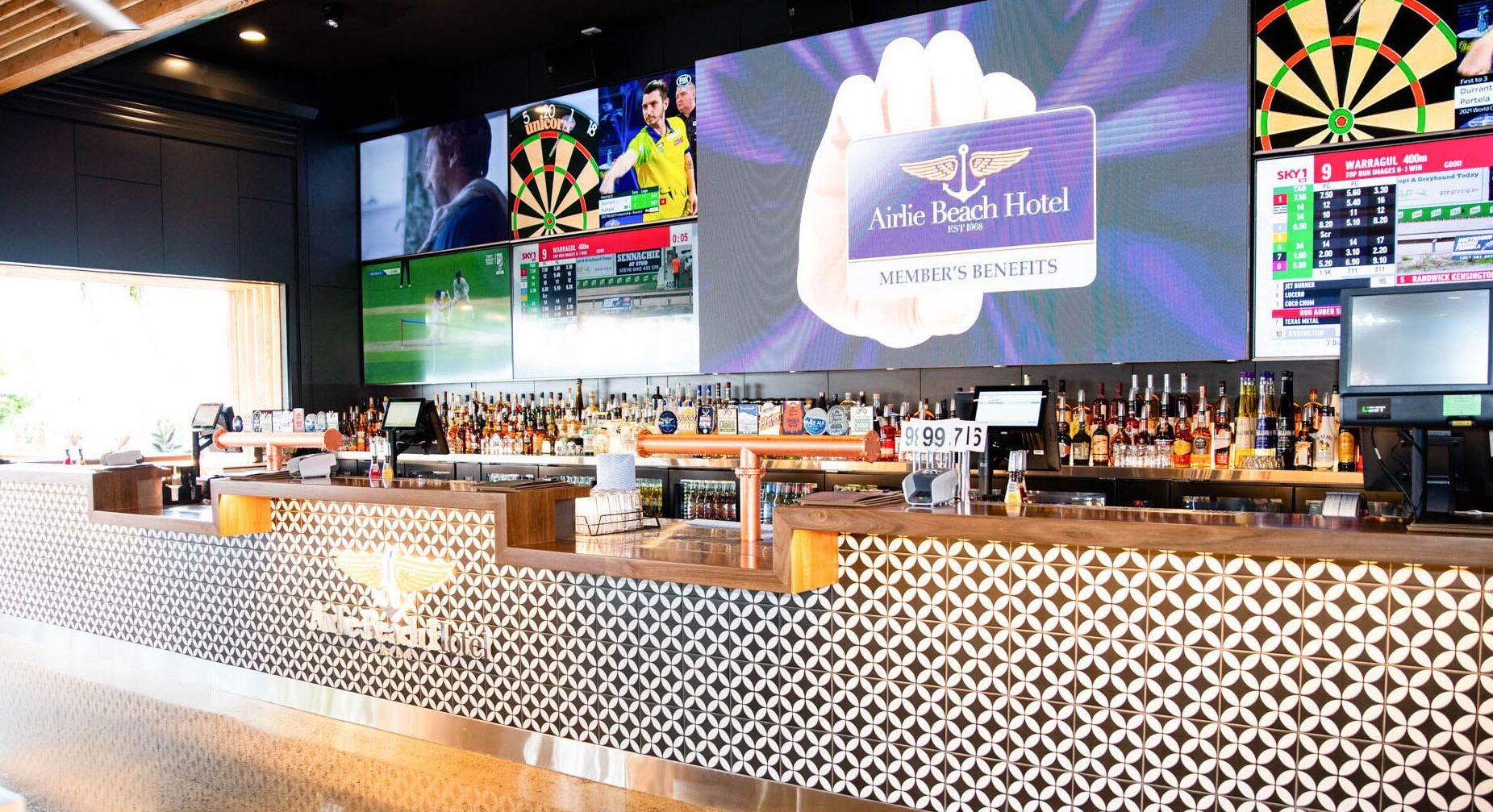 A bar area with a dartboard, a large screen displaying sports, and a sign for Airlie Beach Hotel.