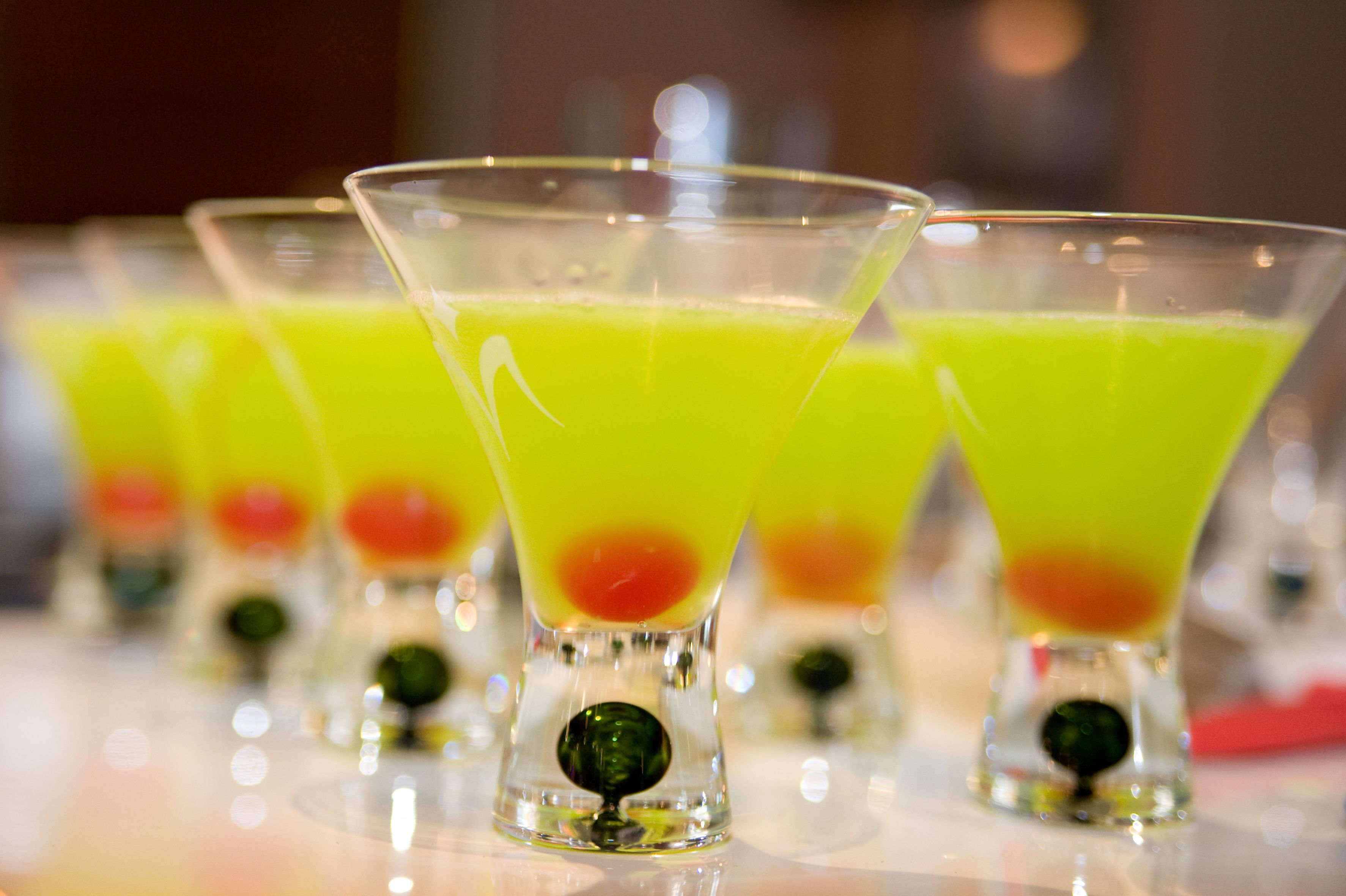 Una fila de copas de martini con líquido verde y una esfera roja en el centro, sobre una superficie reflectante.