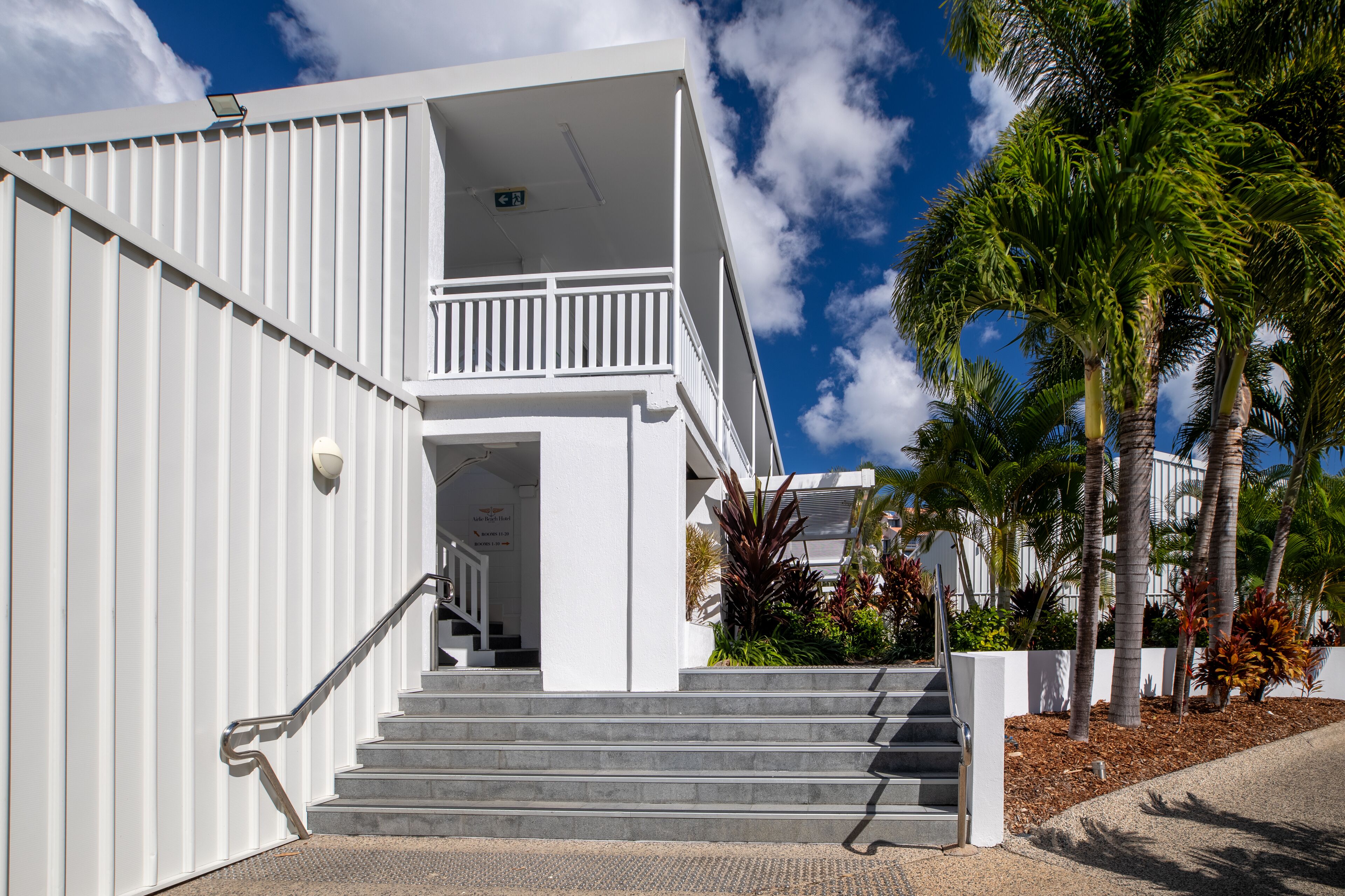 A white building with a staircase leading to a balcony, surrounded by palm trees and tropical plants.