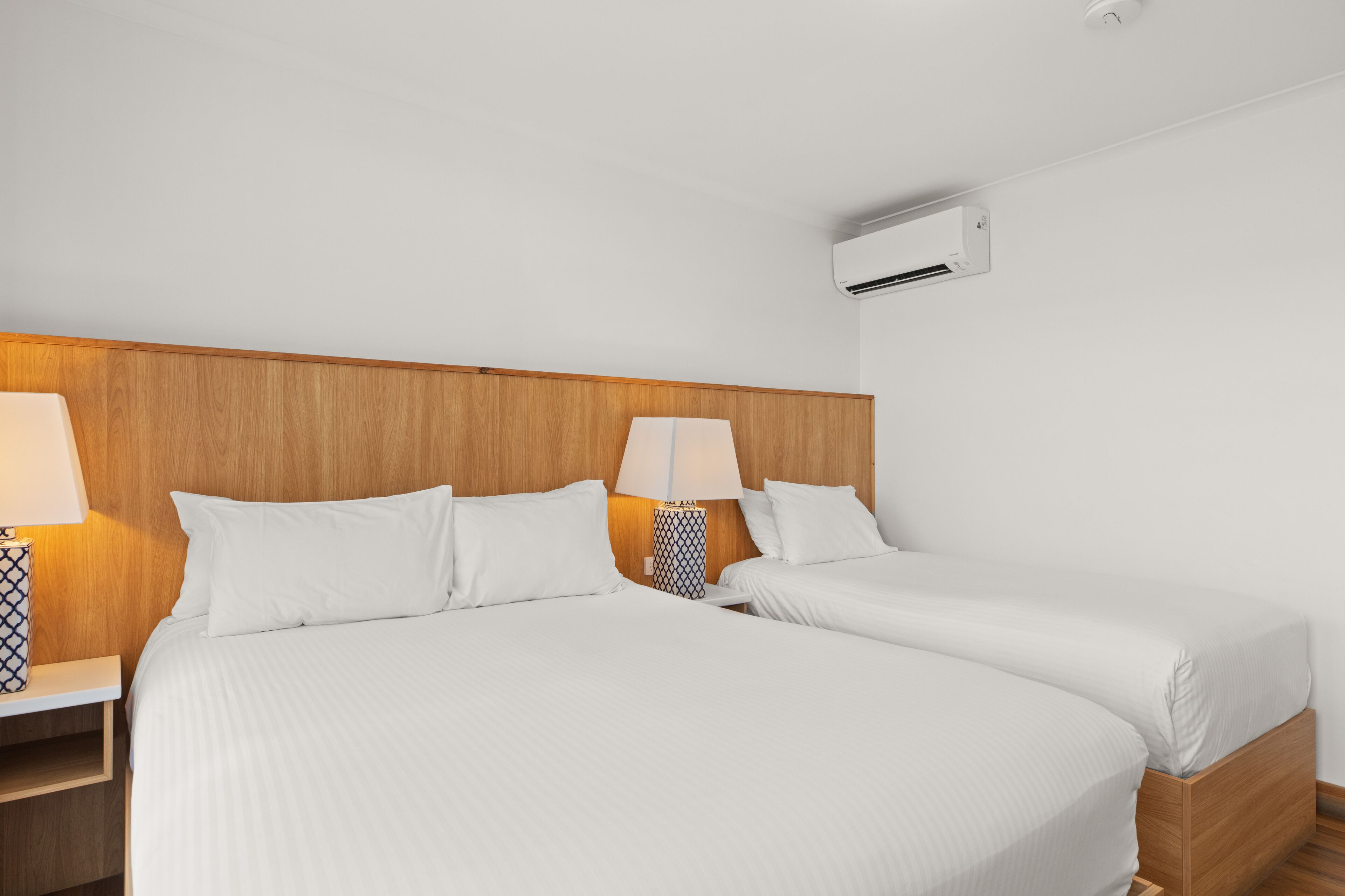 A hotel room with two beds, wooden headboard, white bedding, bedside lamps, and an air conditioning unit.