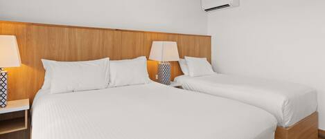 Habitación de hotel con dos camas, cabecero de madera, ropa de cama blanca, lámparas de noche y un equipo de aire acondicionado.