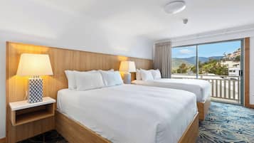 A hotel room with two beds, a wooden headboard, a large window with a view of buildings and hills, and a patterned carpet.