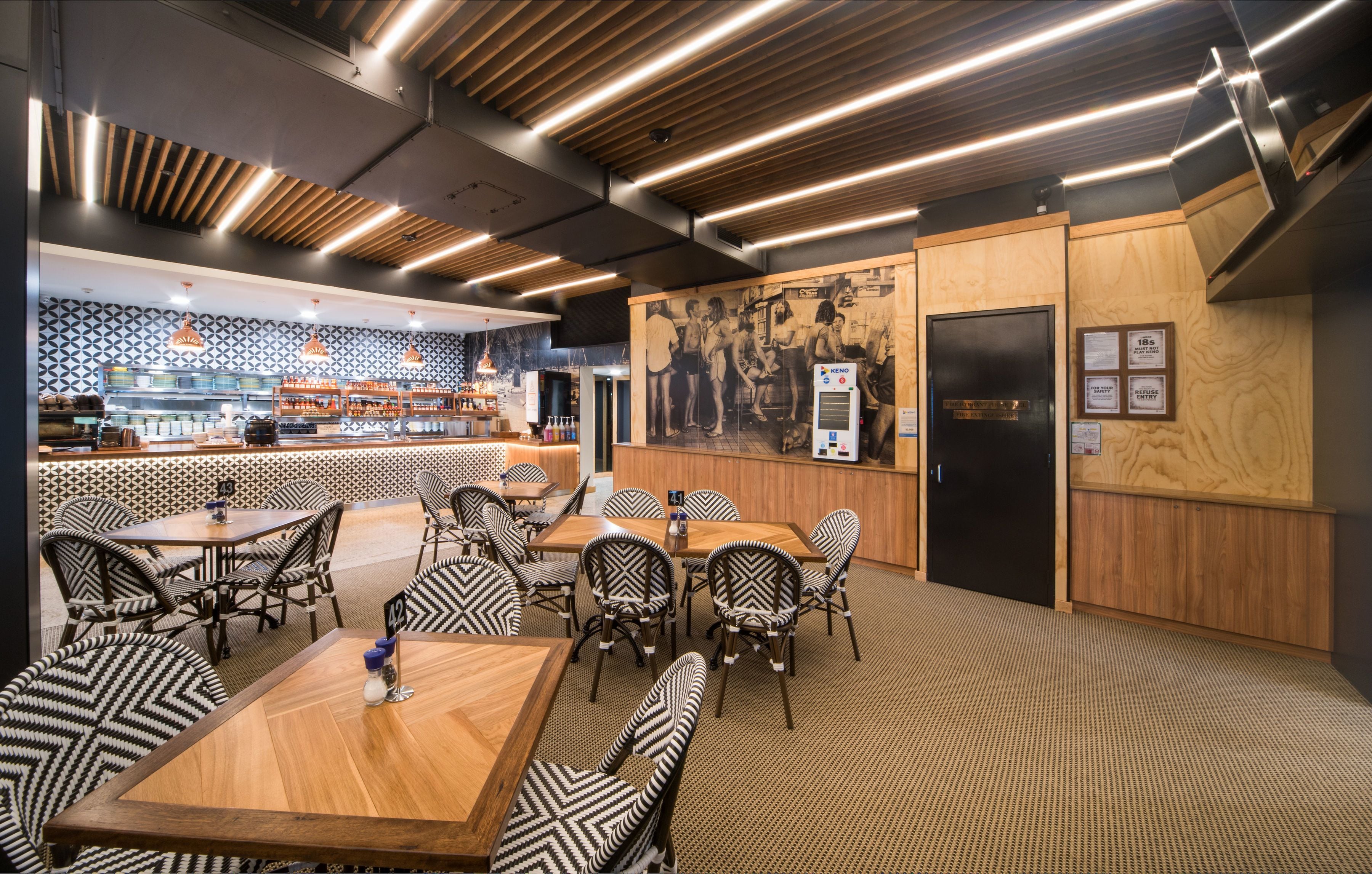 A restaurant with a modern interior, featuring wooden tables and chairs, a patterned wall, and a kitchen area visible in the background.