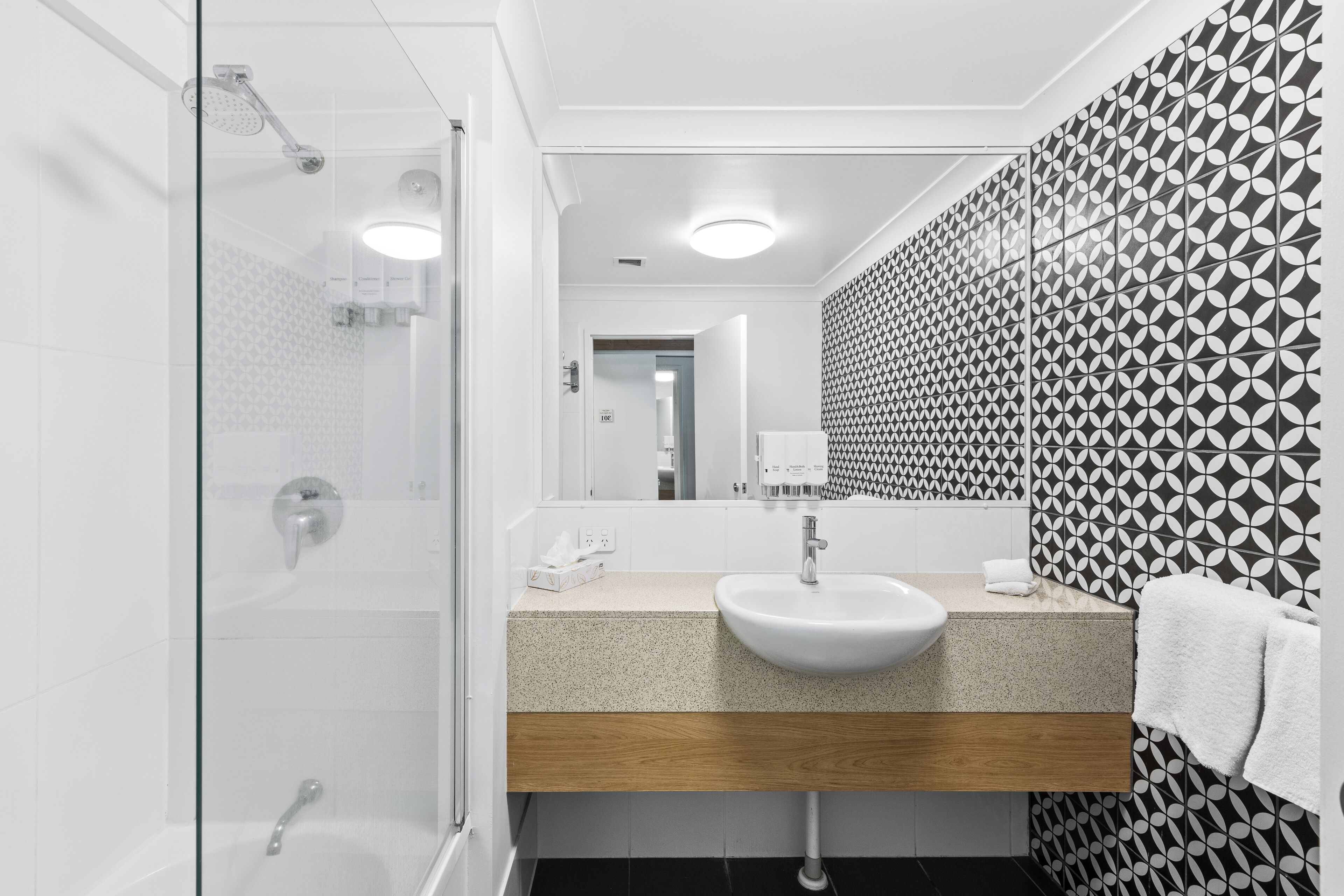 A modern bathroom with a glass shower enclosure, a sink with a wooden countertop, and patterned wallpaper.