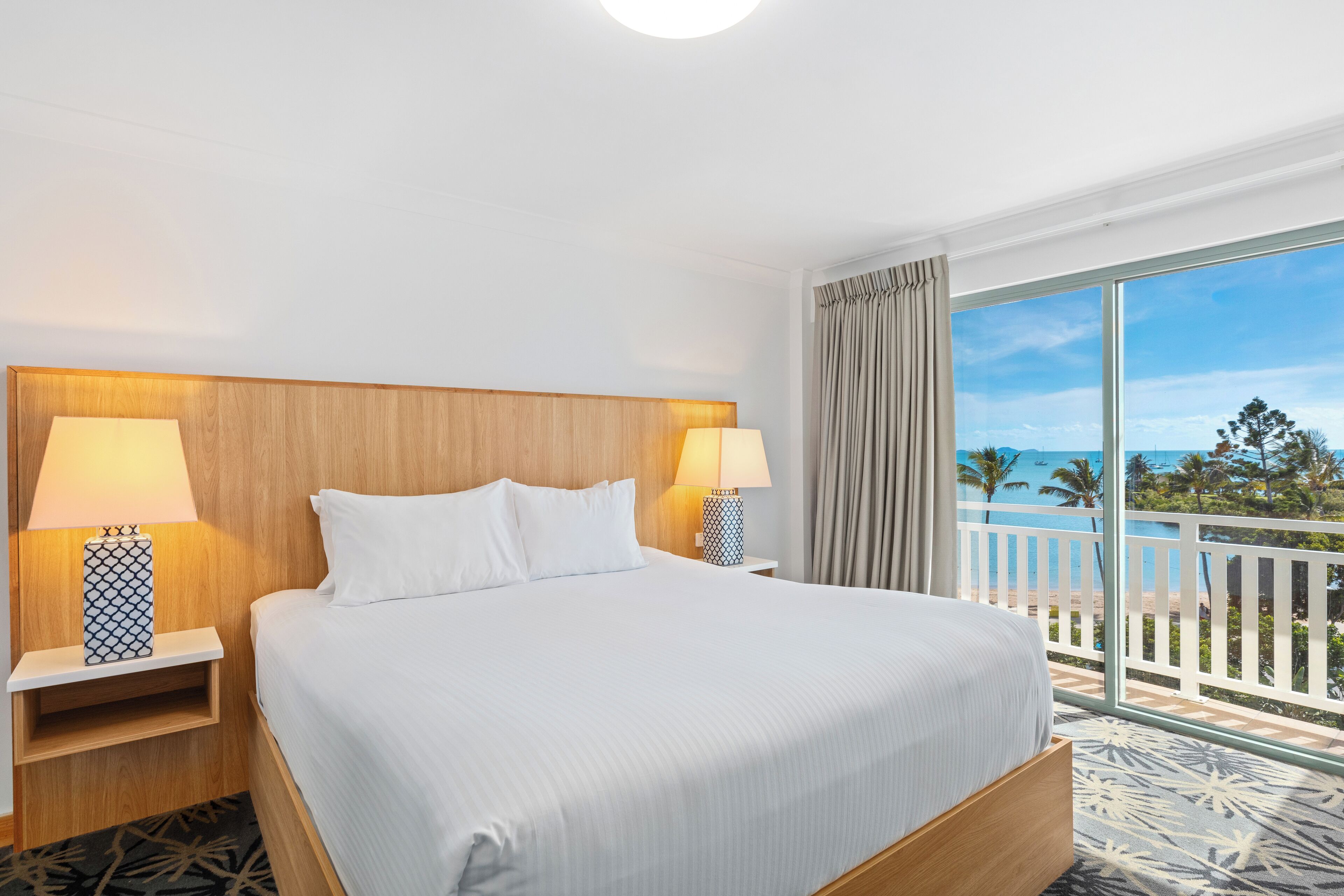 A hotel room with a large bed, two bedside tables with lamps, a view of the beach, and a balcony with a railing.