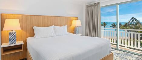 A hotel room with a large bed, two bedside tables with lamps, a view of the beach, and a balcony with a railing.
