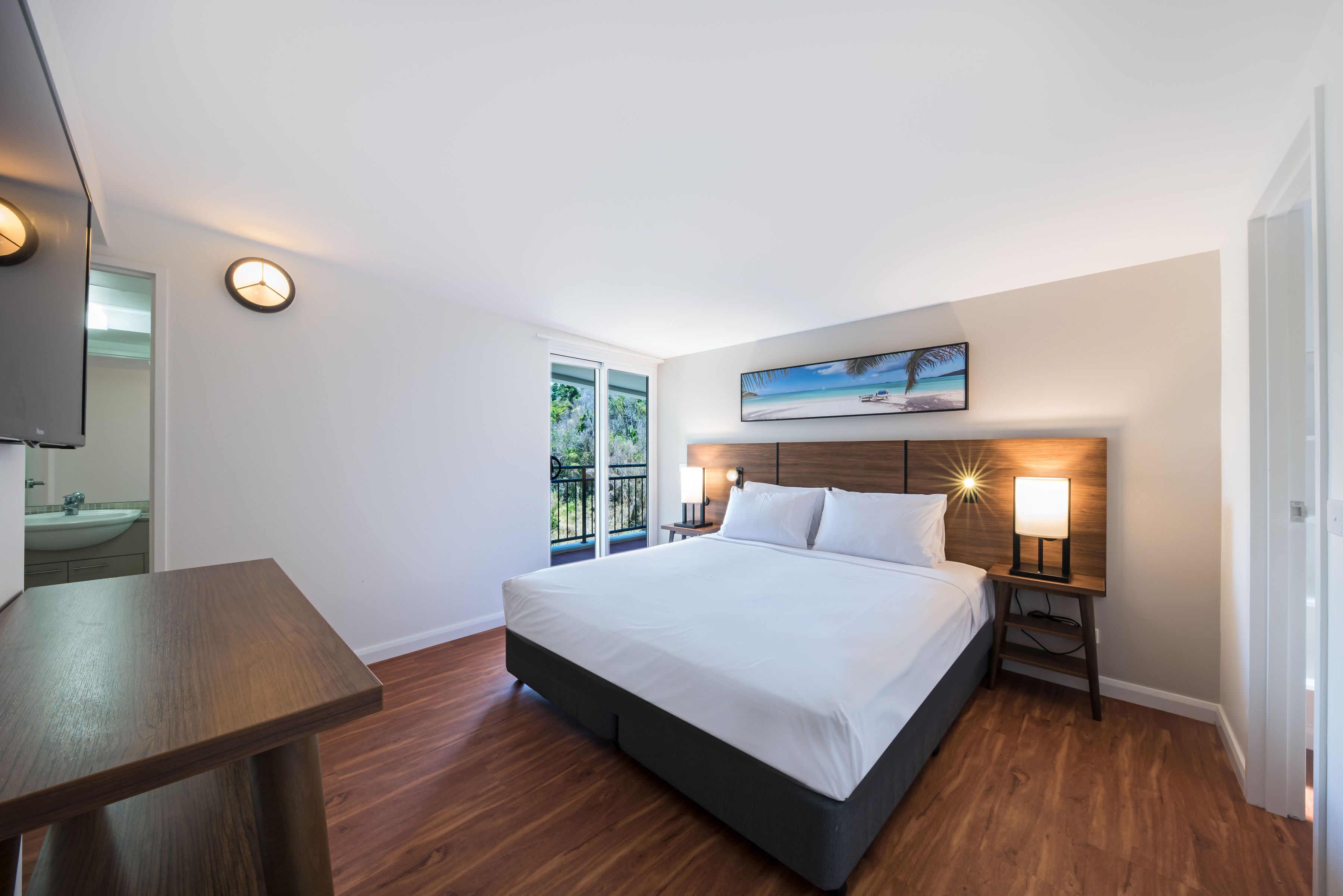 Una camera da letto moderna con un letto grande, un comodino in legno con una lampada, un quadro appeso e vista all'aperto attraverso una finestra.