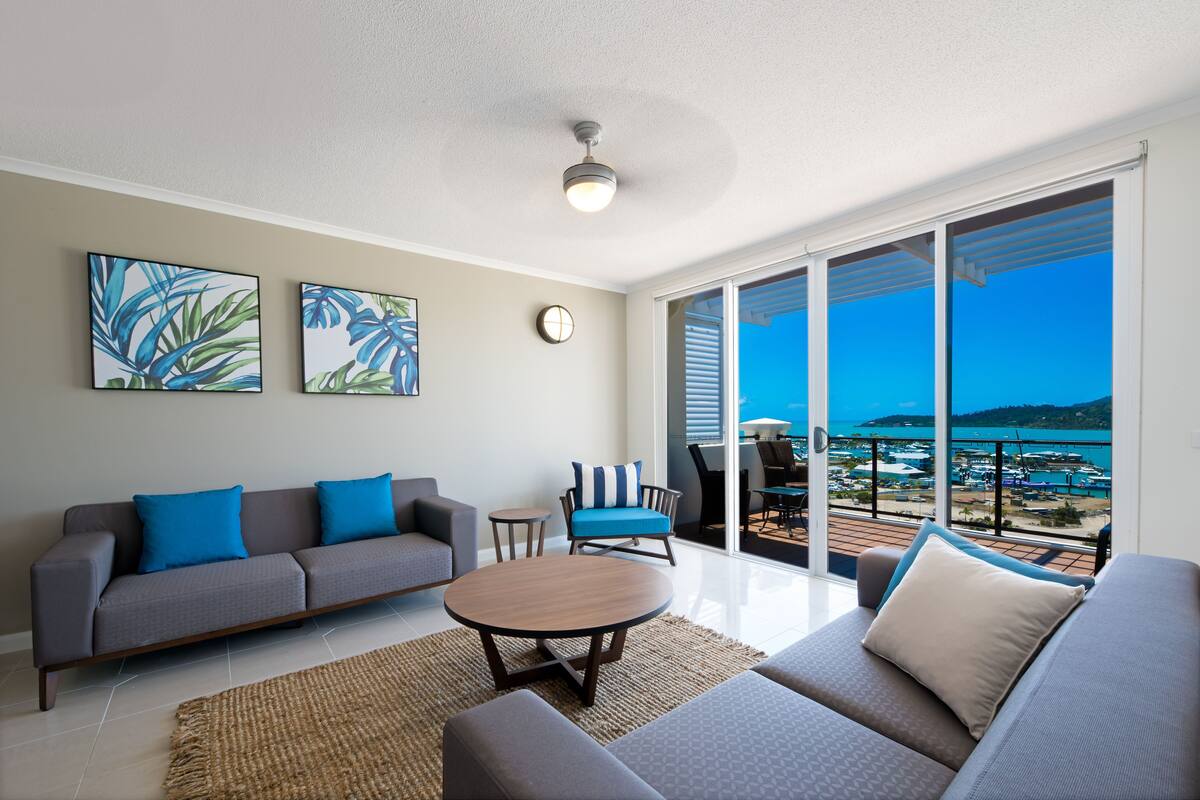 Una sala de estar moderna con sofá, sillón y mesa de centro, un balcón con vista al mar y dos cuadros colgados en la pared.