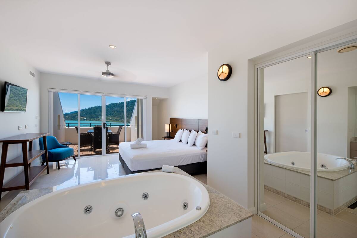 Una habitación de hotel moderna con una cama grande, una zona de estar separada con una silla azul, una bañera grande y un balcón con vistas a las montañas.