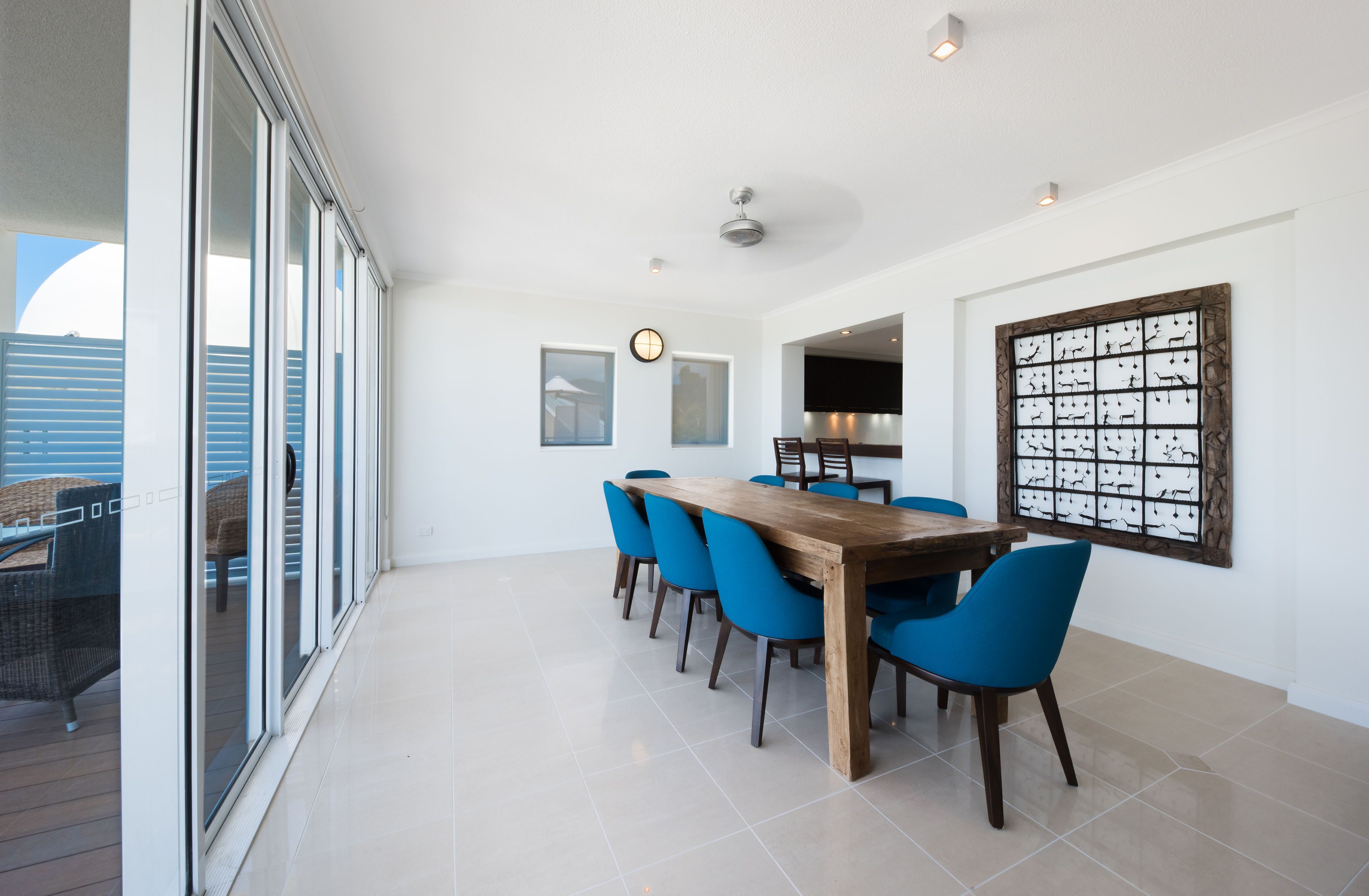 A dining area with a wooden table and blue chairs, large windows, and a decorative wall piece.